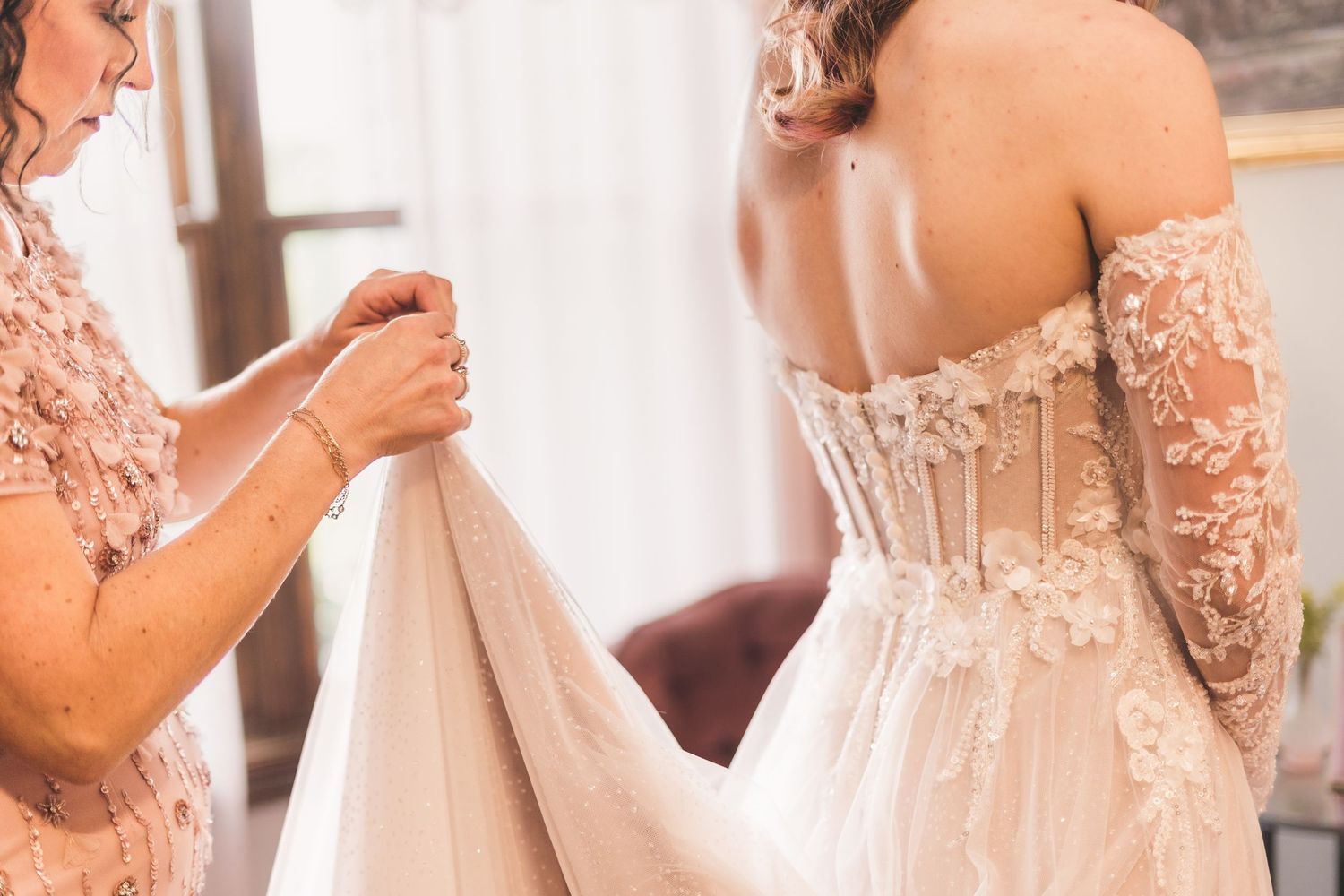 Bride in lace corset wedding gown being helped with dress by bridesmaid in floral pink top.