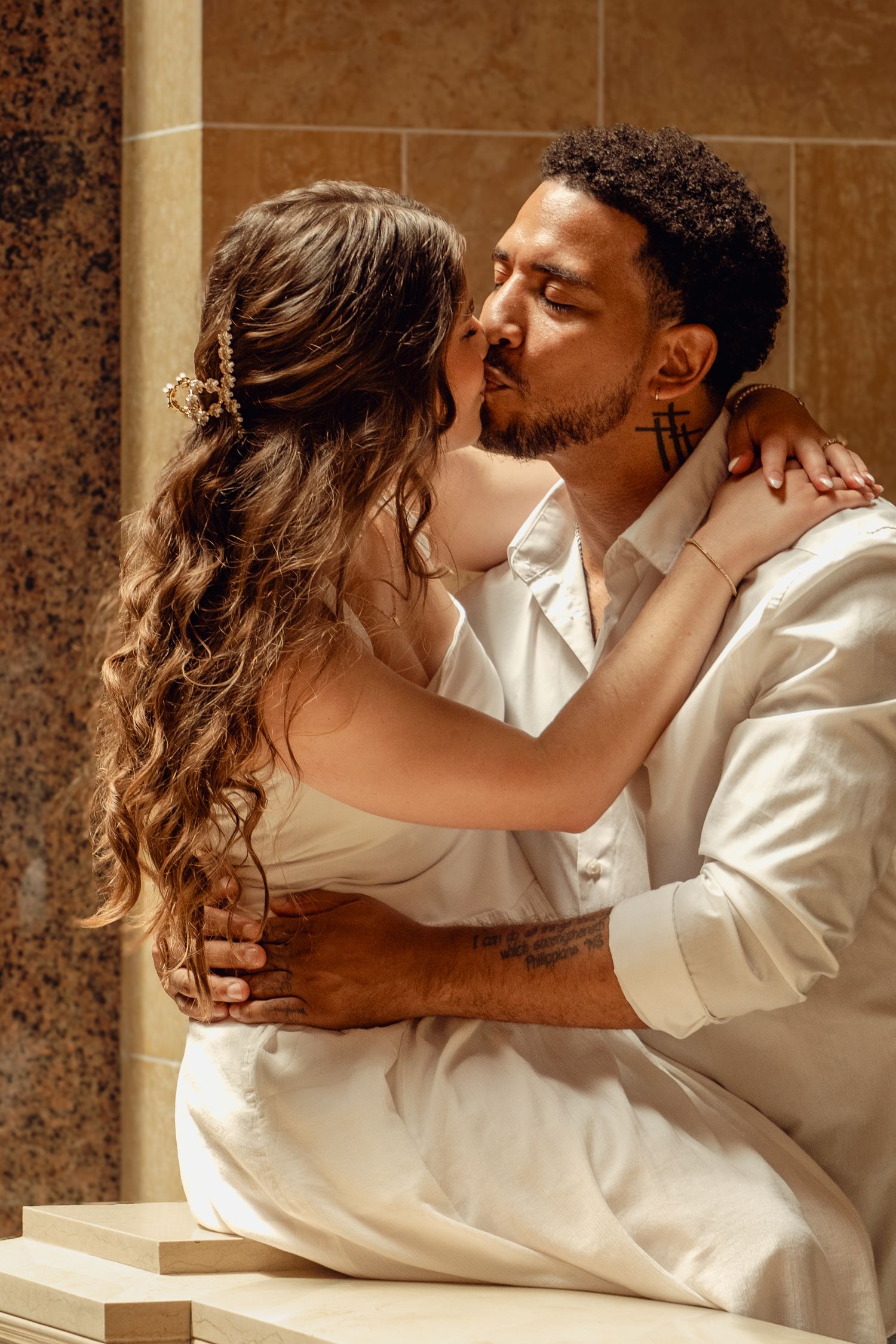 A couple in white outfits sharing a romantic kiss, with the woman's arms around the man's neck in an elegant setting.