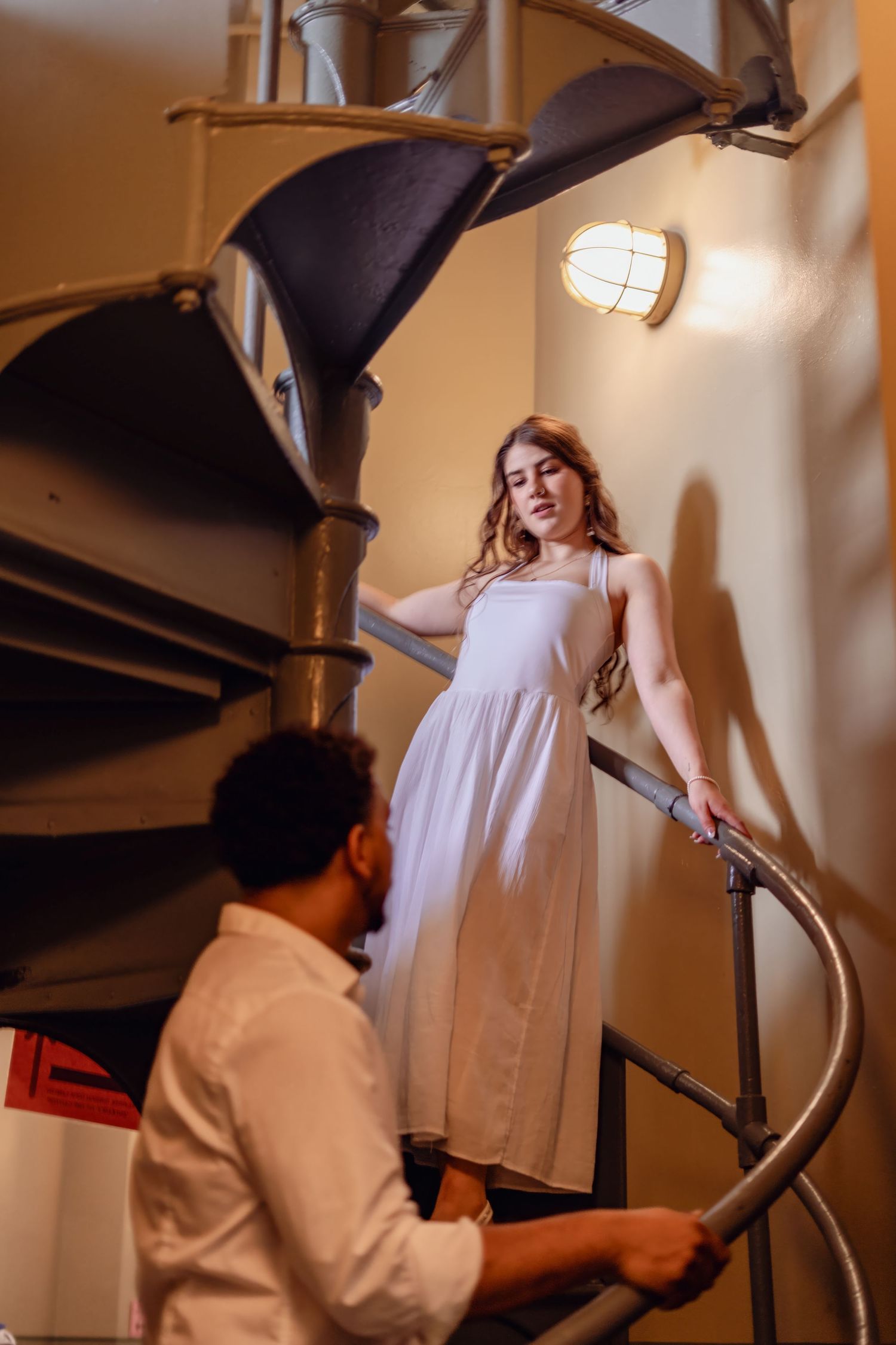 Elegant woman in white dress on spiral staircase looking down at man in white shirt below.