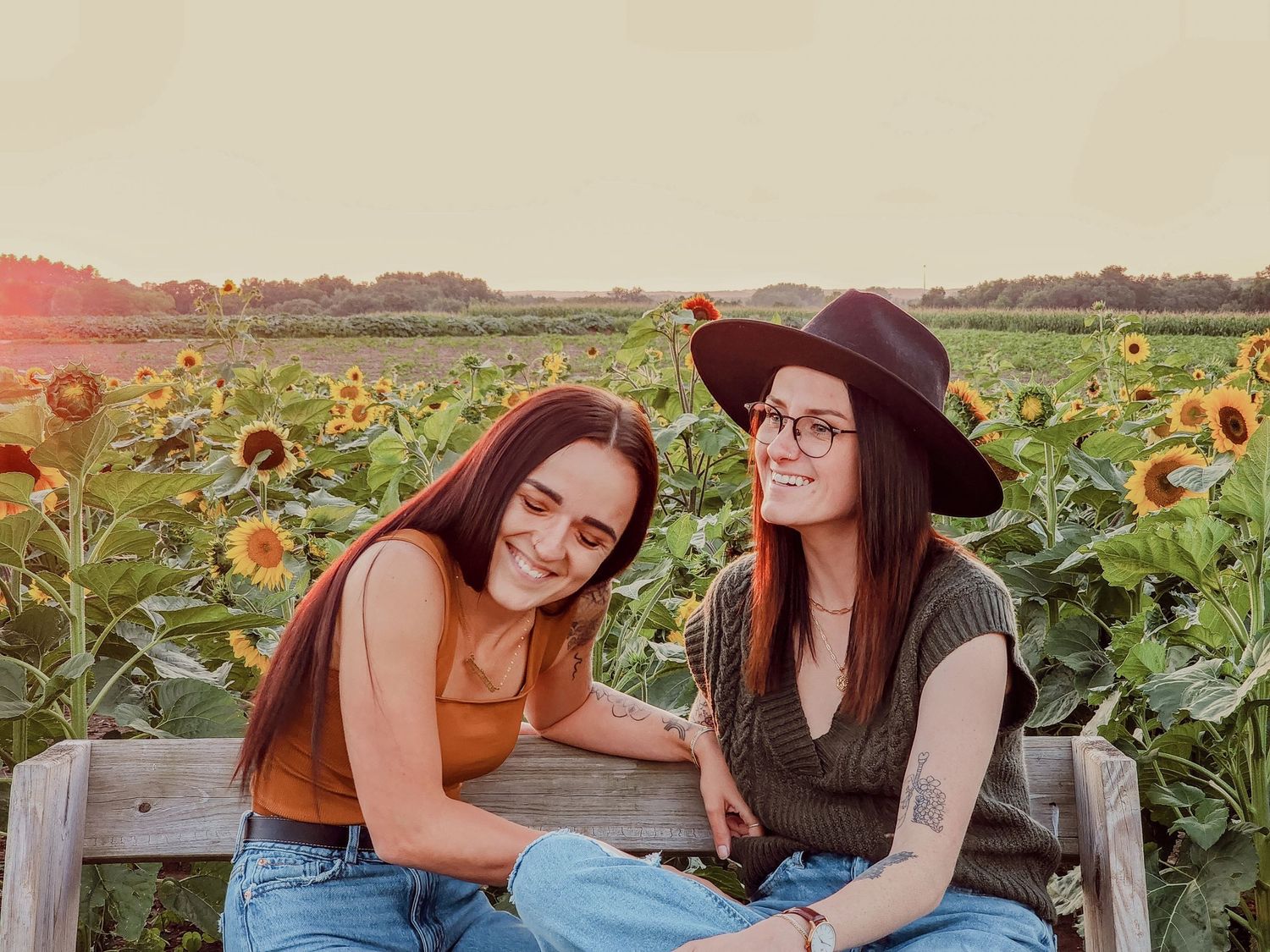 Friends share a laugh while sitting on a bench in a sunflower field at sunset.
