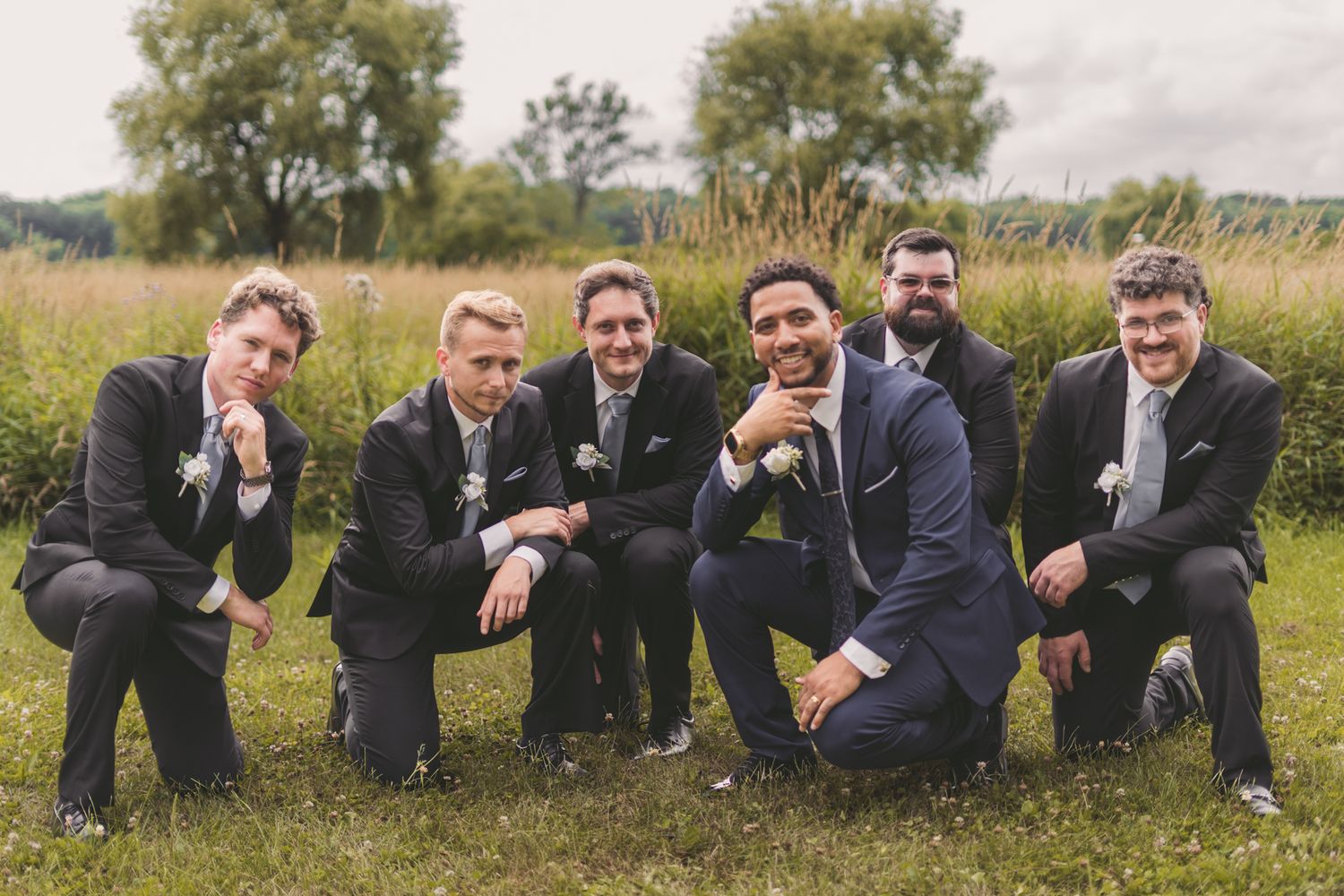 Six groomsmen in navy suits crouching and posing together outdoors in a green field at a wedding.