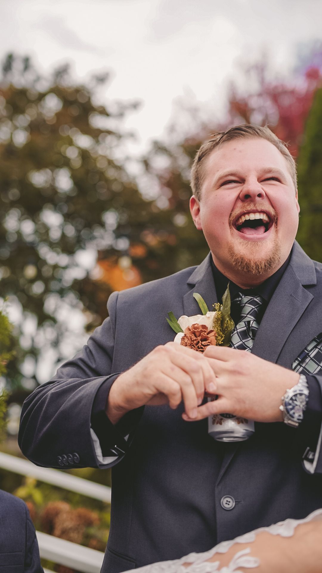 Groom in grey suit laughing while eating during outdoor wedding reception.