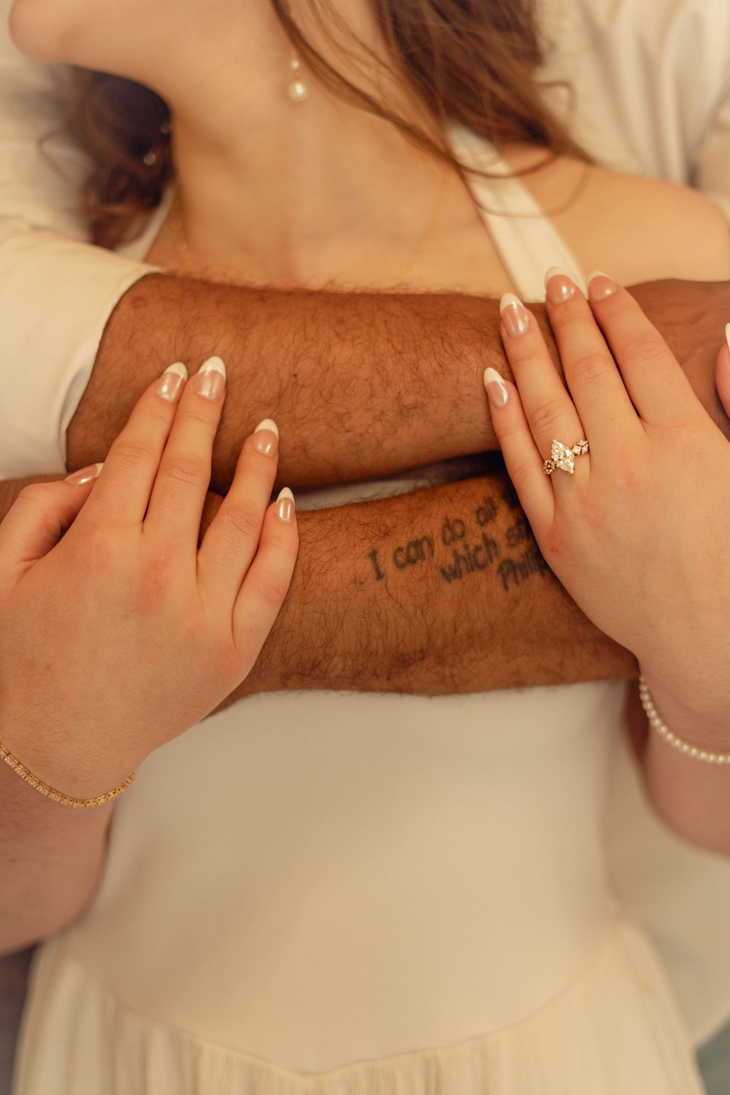 Close-up of engaged couple holding hands, woman wearing diamond ring and pearl bracelet, man with arm tattoo.