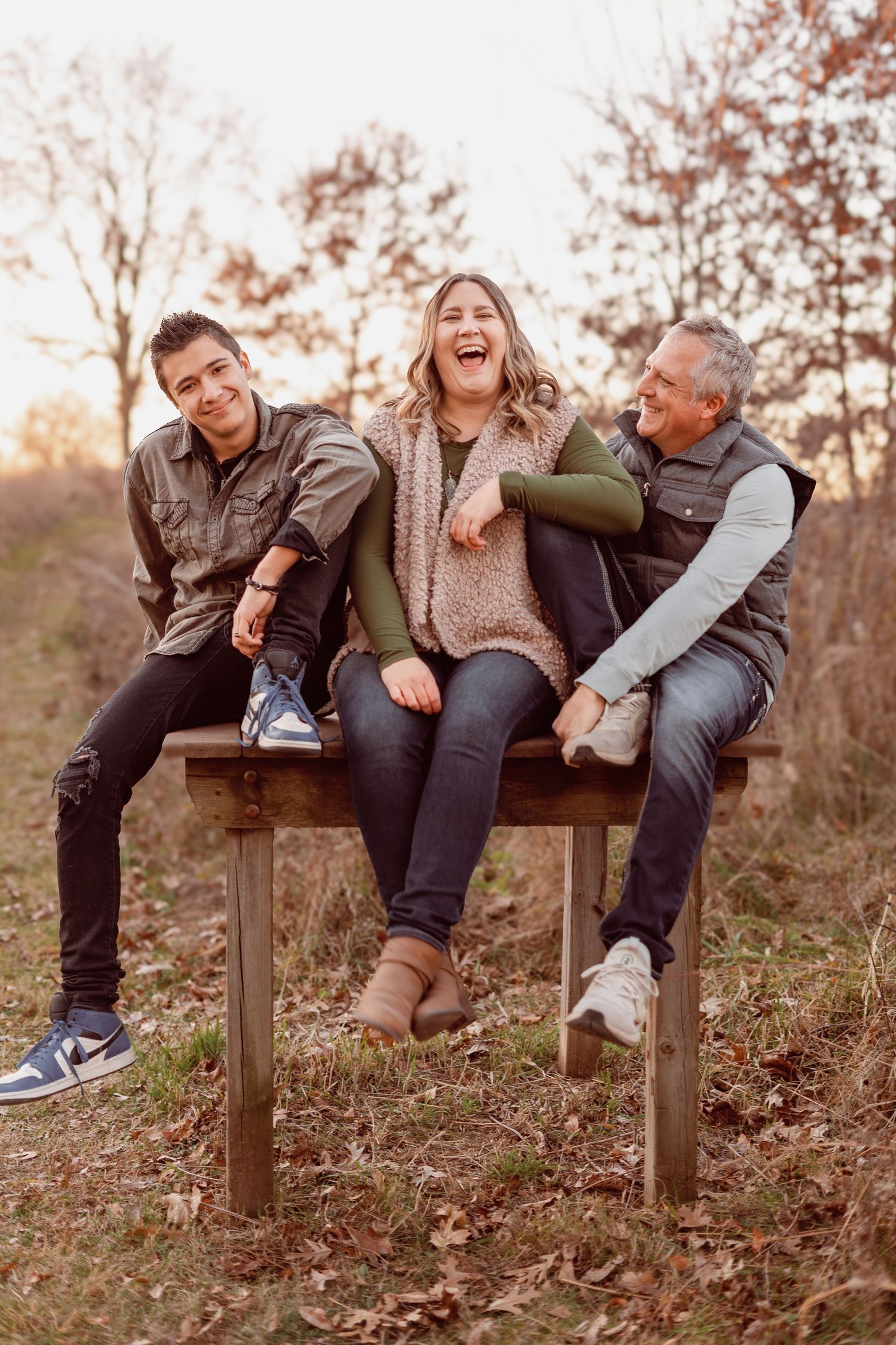 Family of three laughing together on a wooden bench outdoors during autumn, surrounded by fall foliage.