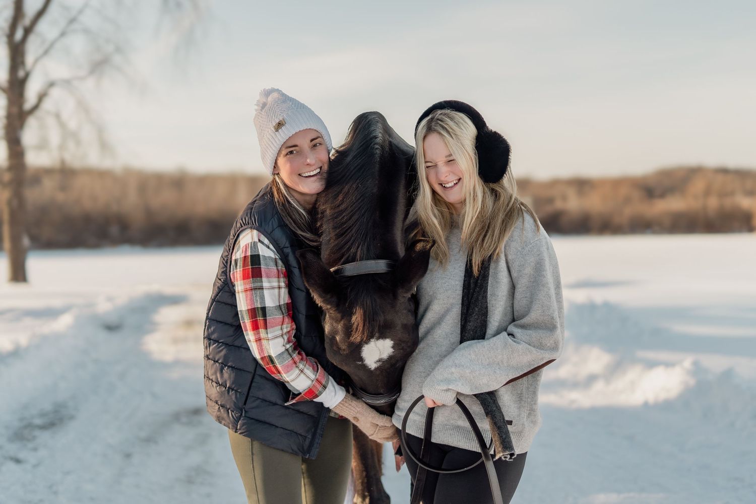 Two smiling women in winter outfits cuddle a dark horse in a snowy outdoor setting on a sunny day.