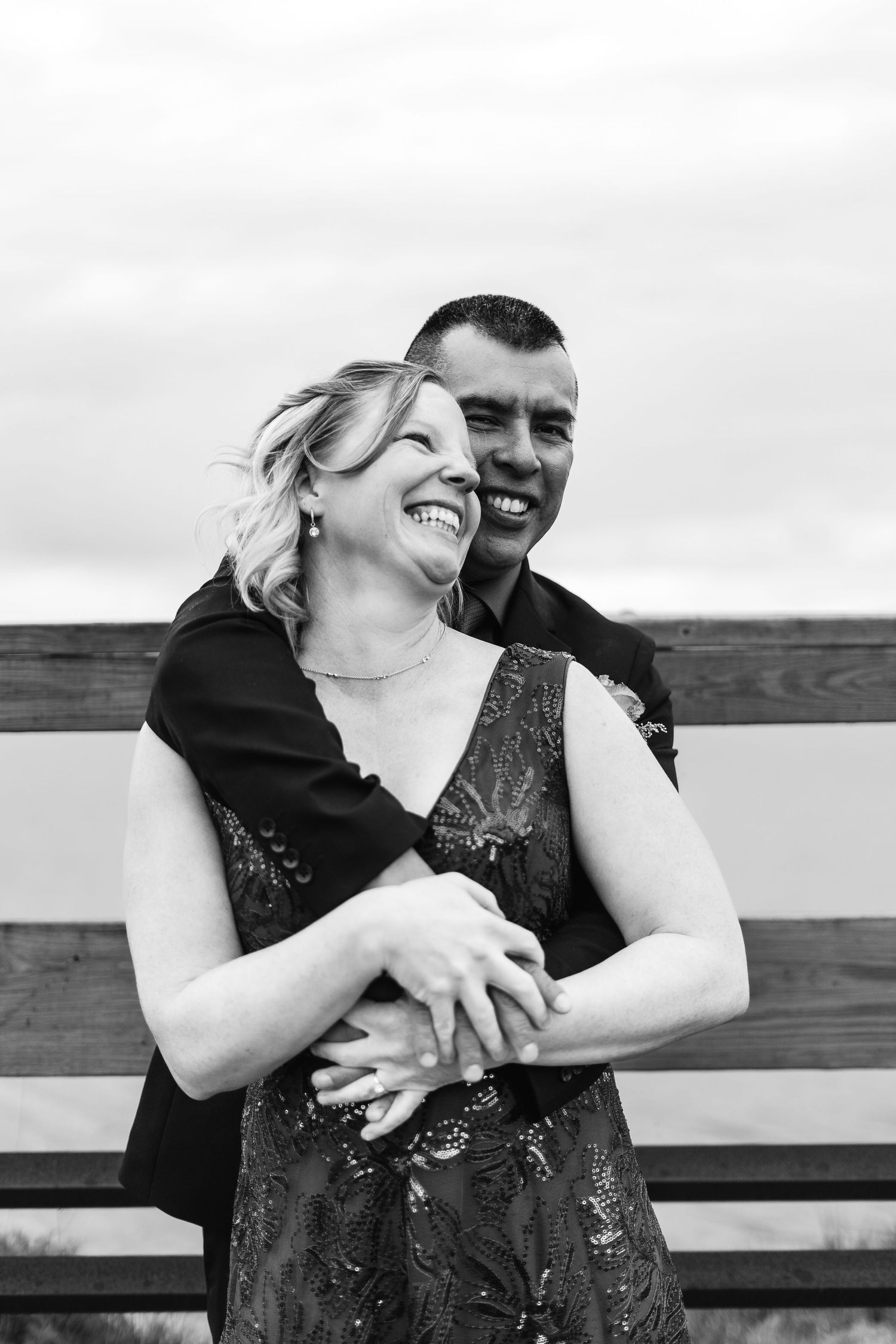 Black and white photo of a laughing couple embracing outdoors near a wooden fence, sharing a joyful romantic moment.