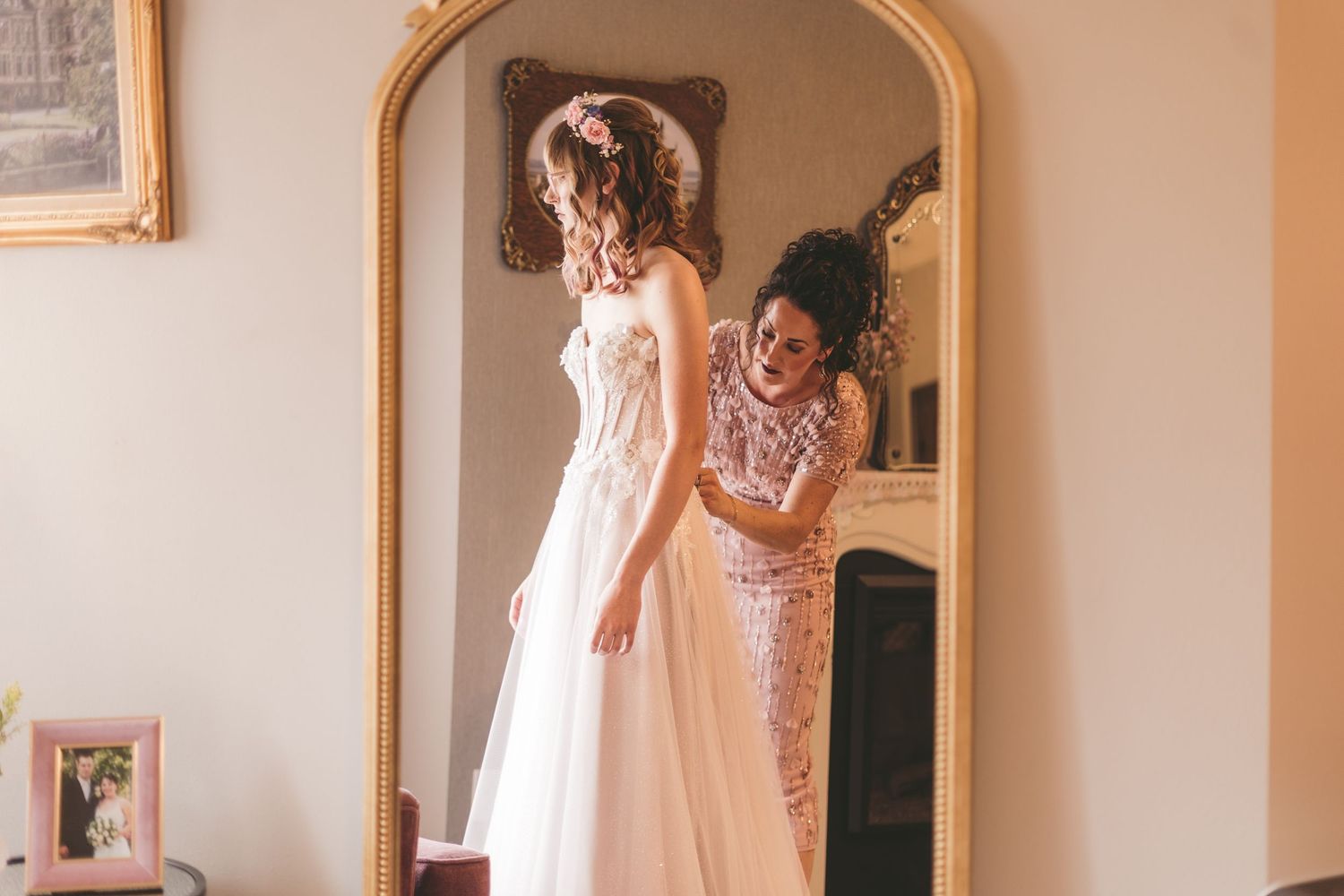 Bridesmaid in sequin dress helping bride into white wedding gown in front of ornate gold mirror.
