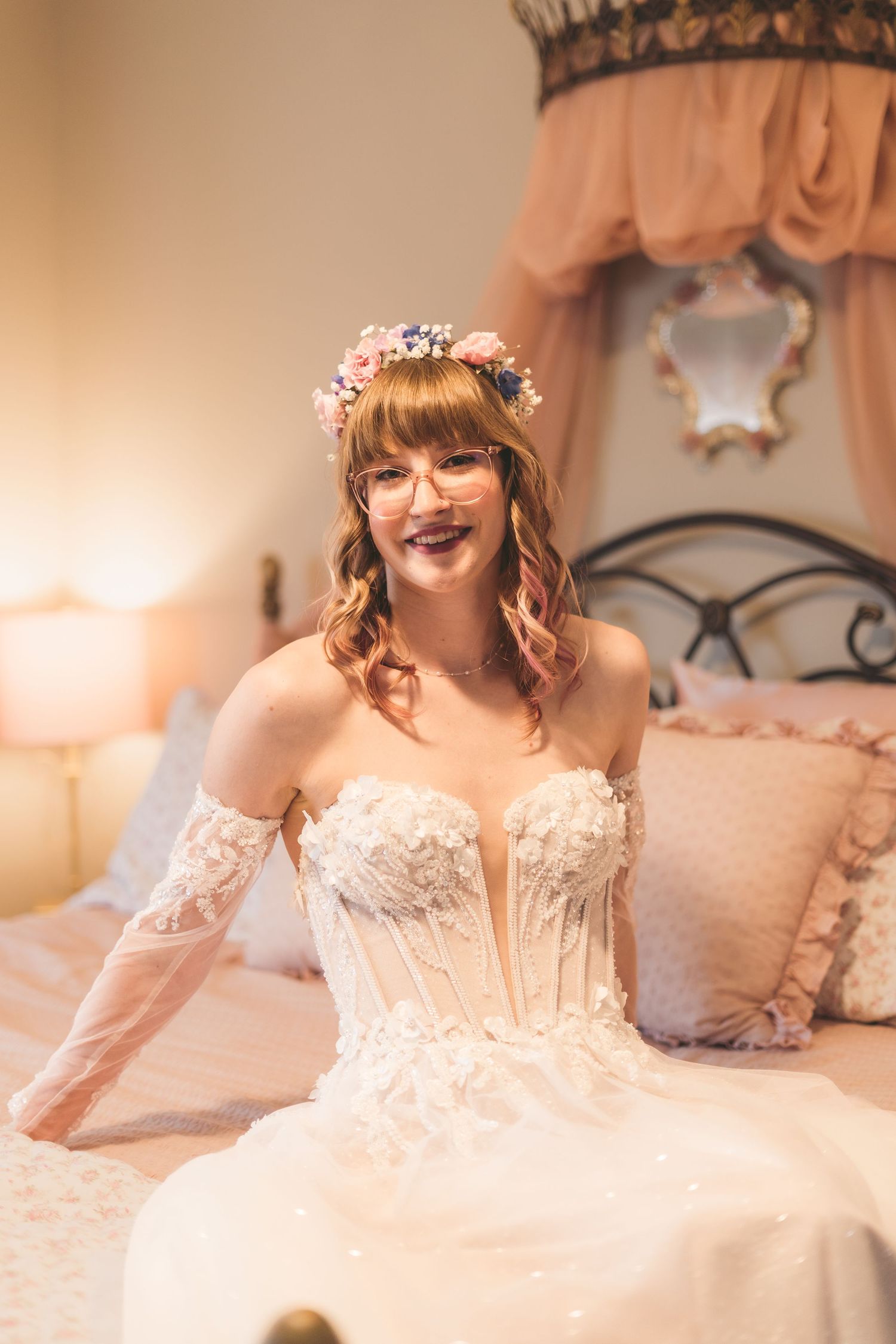 Smiling bride in white corset gown with floral crown and glasses seated on bed.