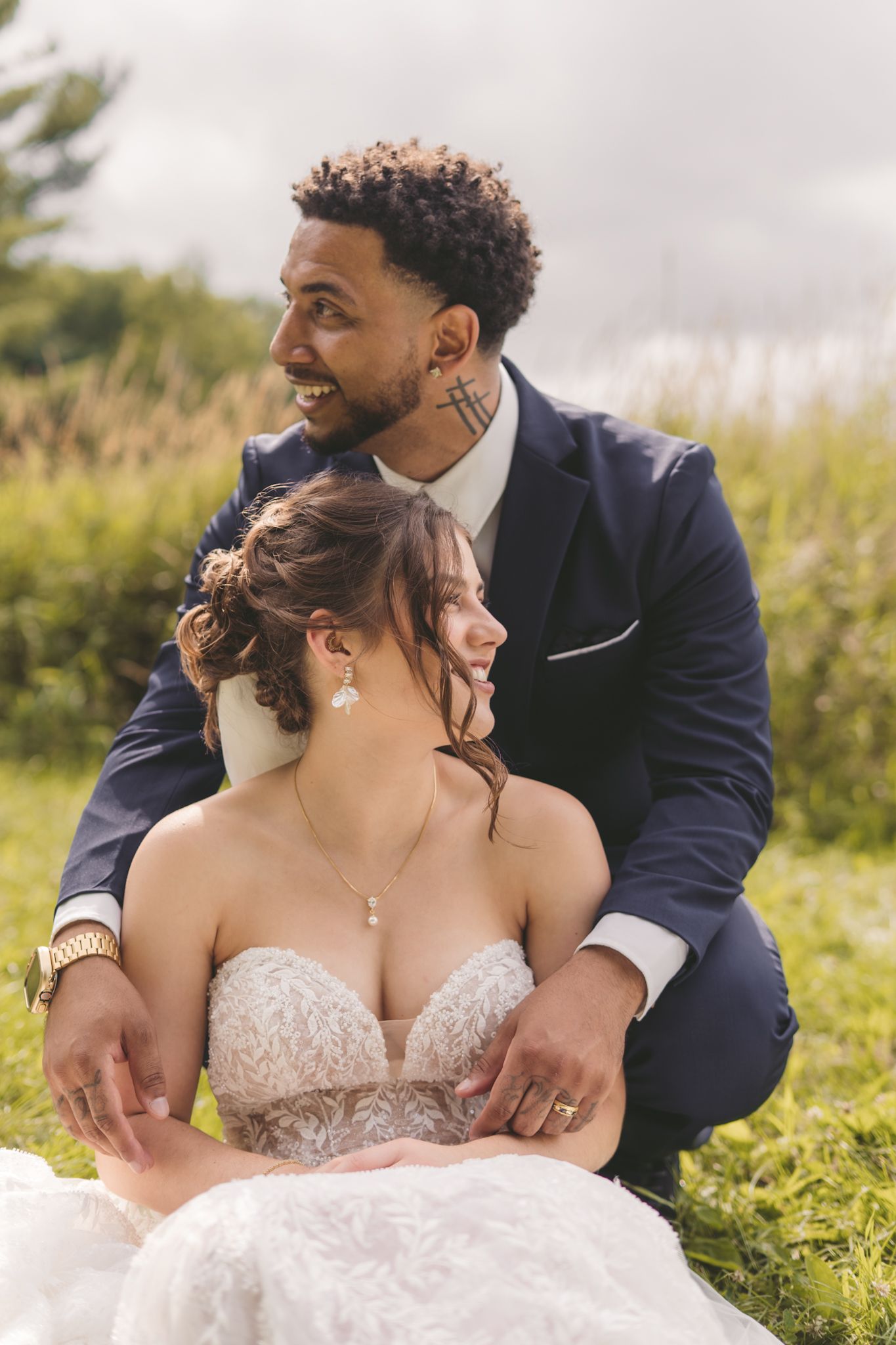 Bride and groom sitting outdoors on grass, groom in navy suit smiling, bride in strapless lace gown on their wedding day.