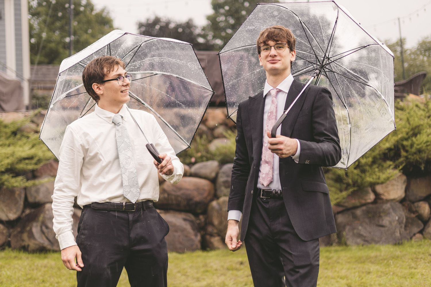 Two young men in suits holding clear umbrellas outdoors on a rainy day at a formal event.
