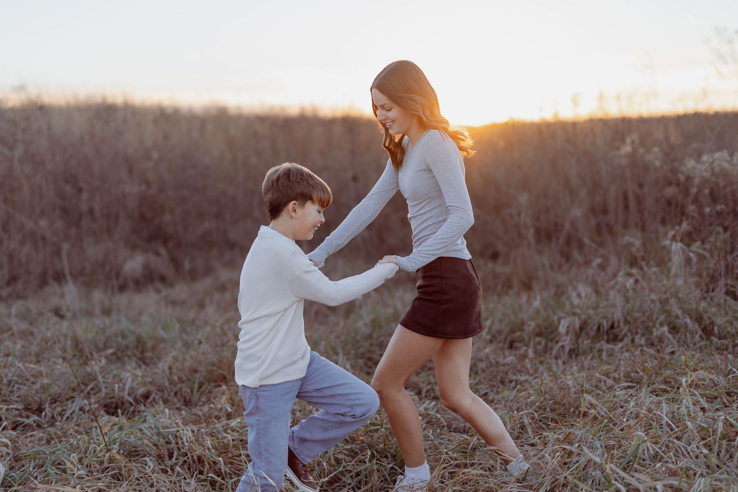 A boy kneels holding hands with a teenage girl in a sunny field at golden hour.