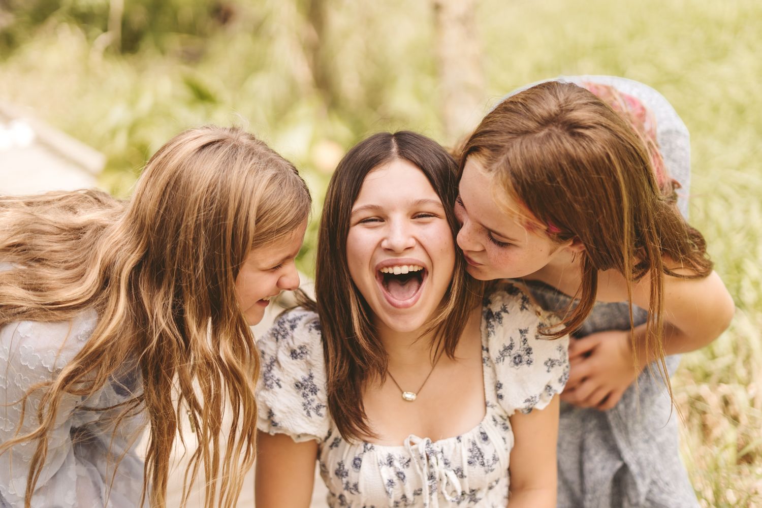 Three laughing girls hugging outdoors, sharing a joyful moment together in a sunny green setting.