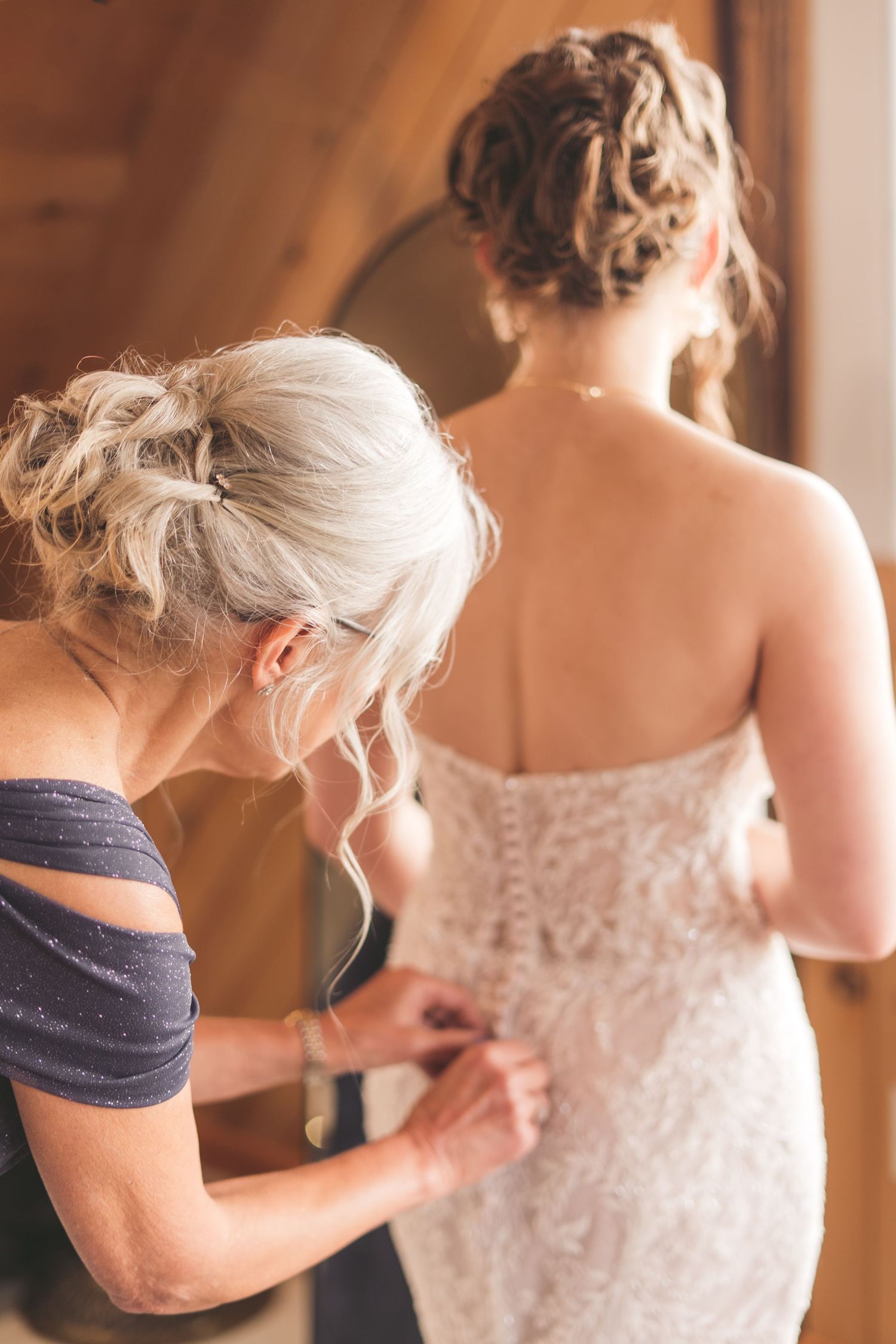 Woman in blue dress buttoning the back of a bride's lace wedding gown in a warm wooden venue.
