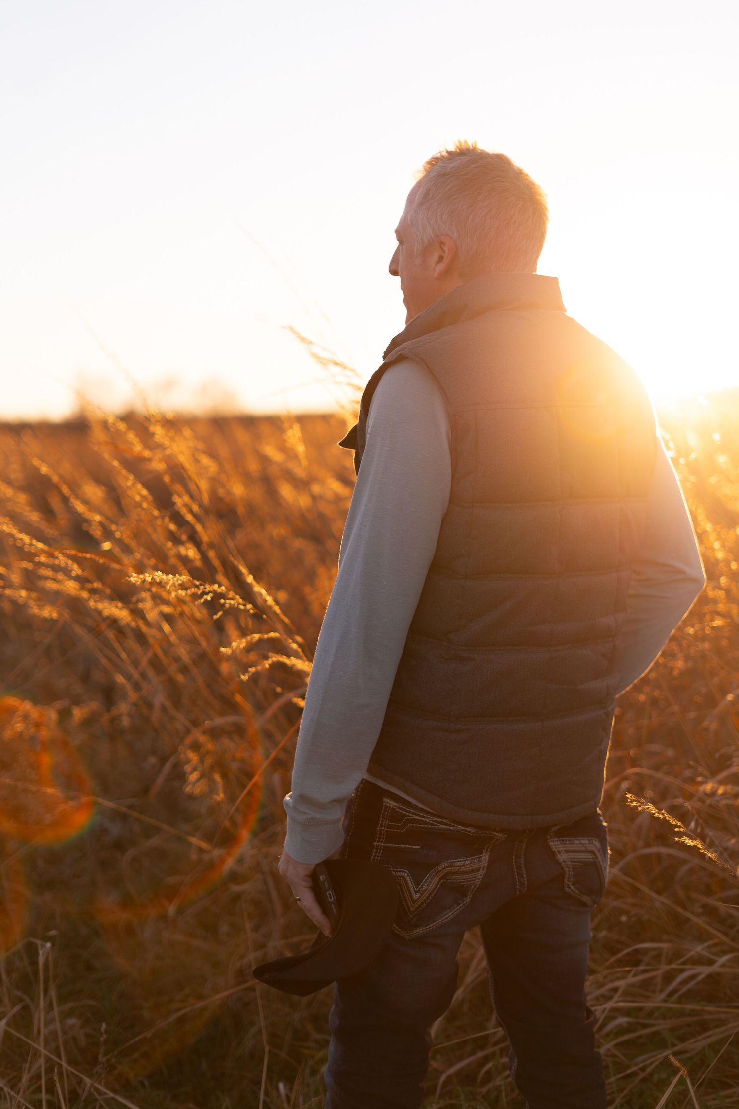 Man in vest standing in golden wheat field at sunset, backlit by warm sunlight in autumn landscape.