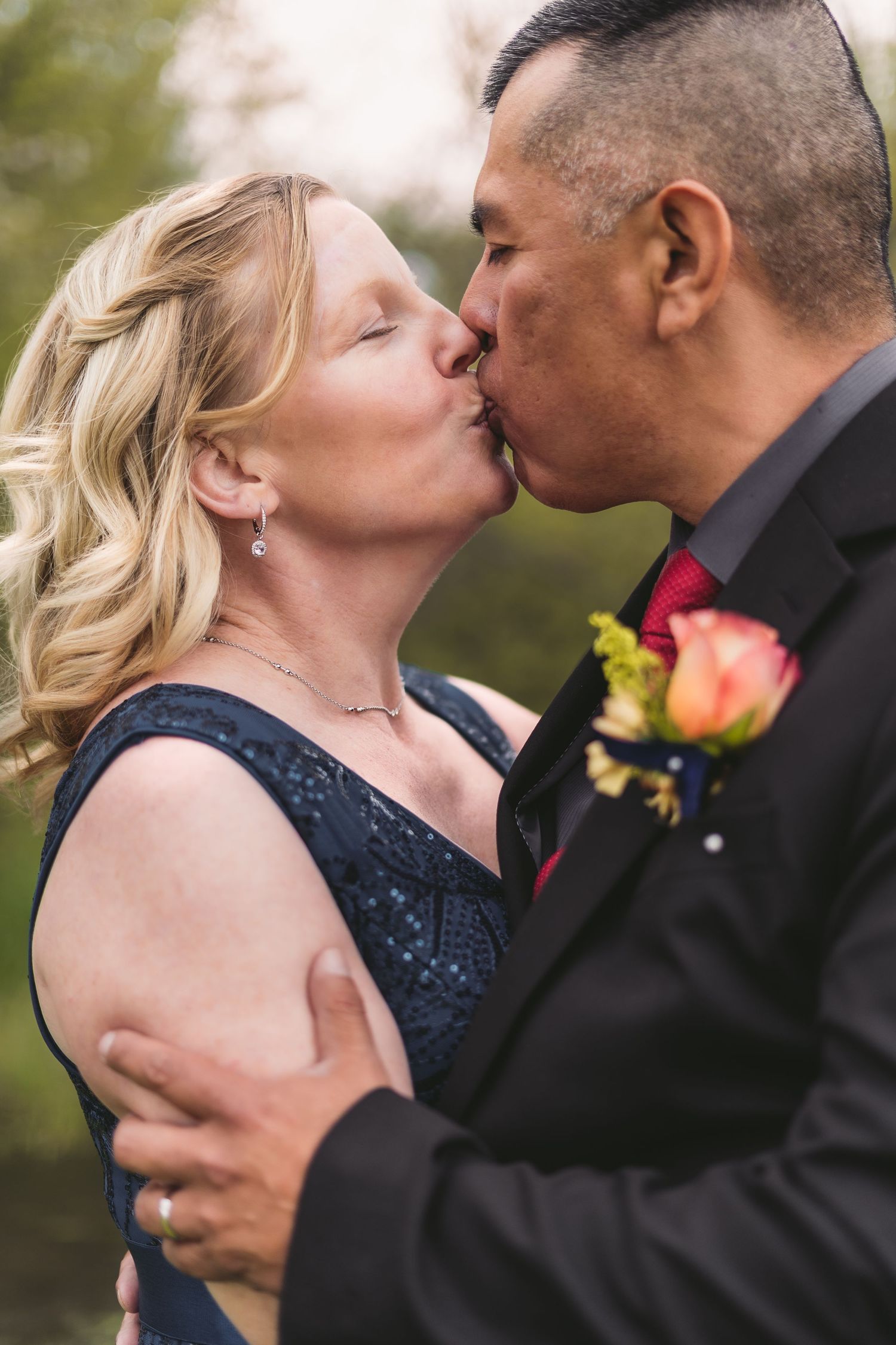 Couple sharing a romantic kiss at their wedding, man in black suit with boutonniere, woman in navy blue dress.