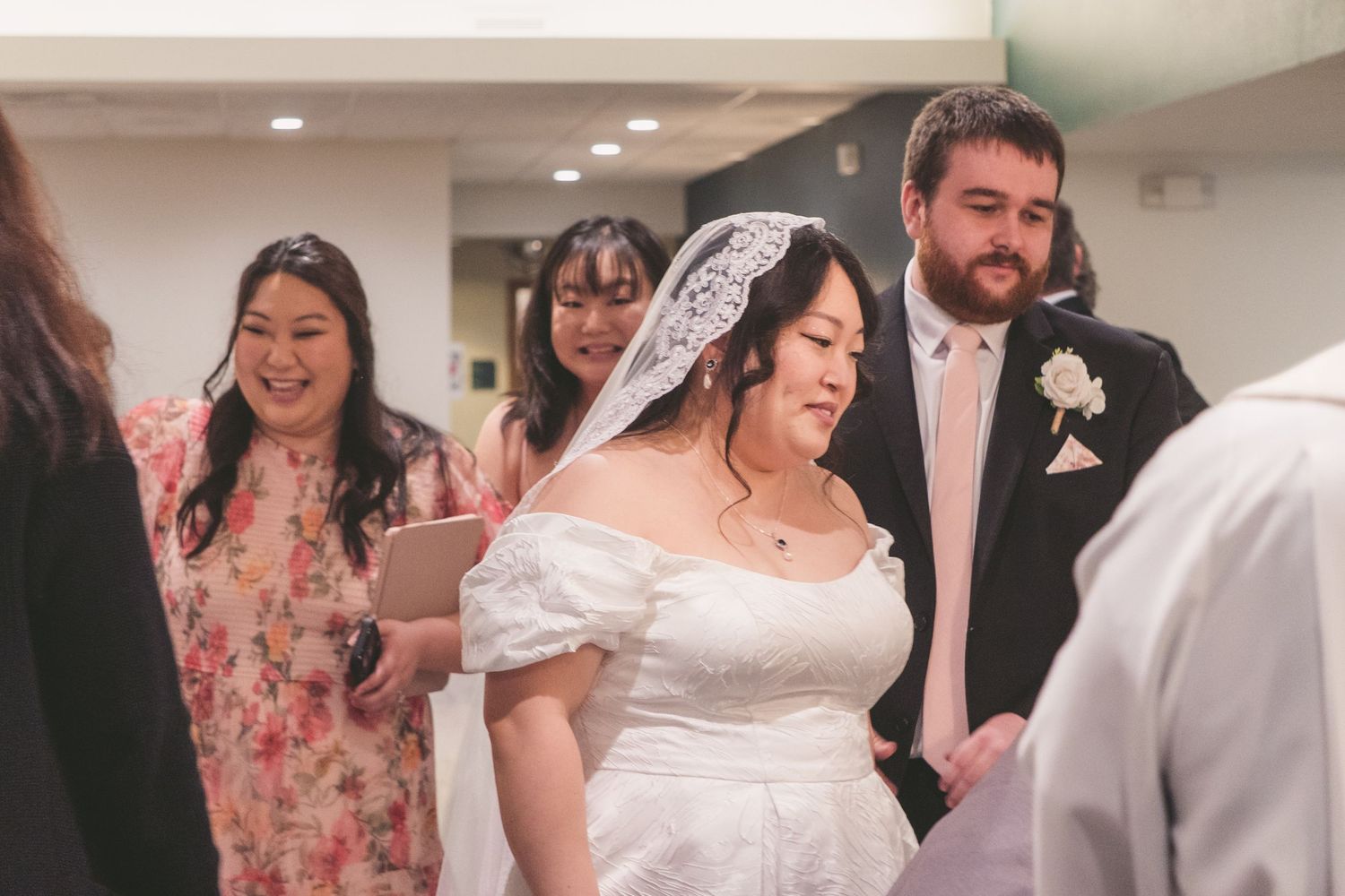 Bride in white wedding dress and veil stands with groom in pink tie during wedding ceremony with guests smiling behind.