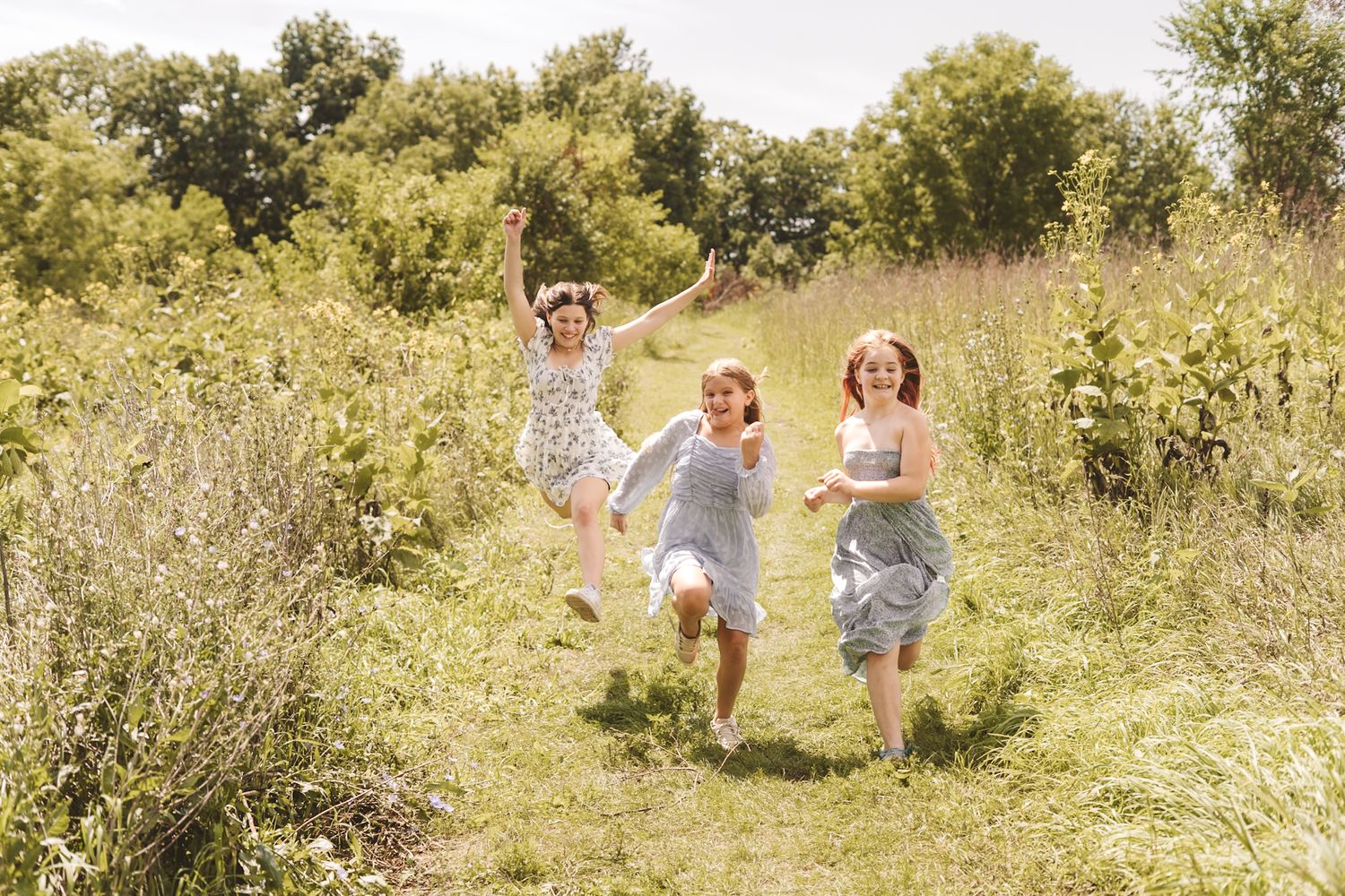 Three girls in floral dresses running joyfully through a sunny green meadow path surrounded by lush trees.