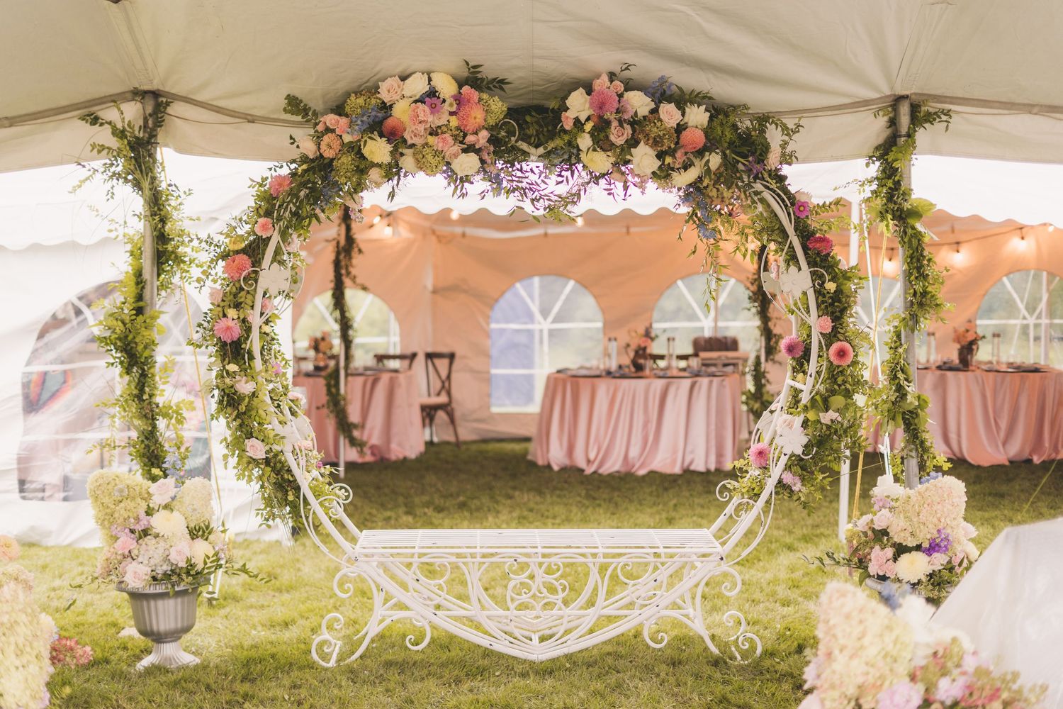 Elegant outdoor wedding tent with floral arch, pink roses, greenery, and blush pink draped tables on green grass.