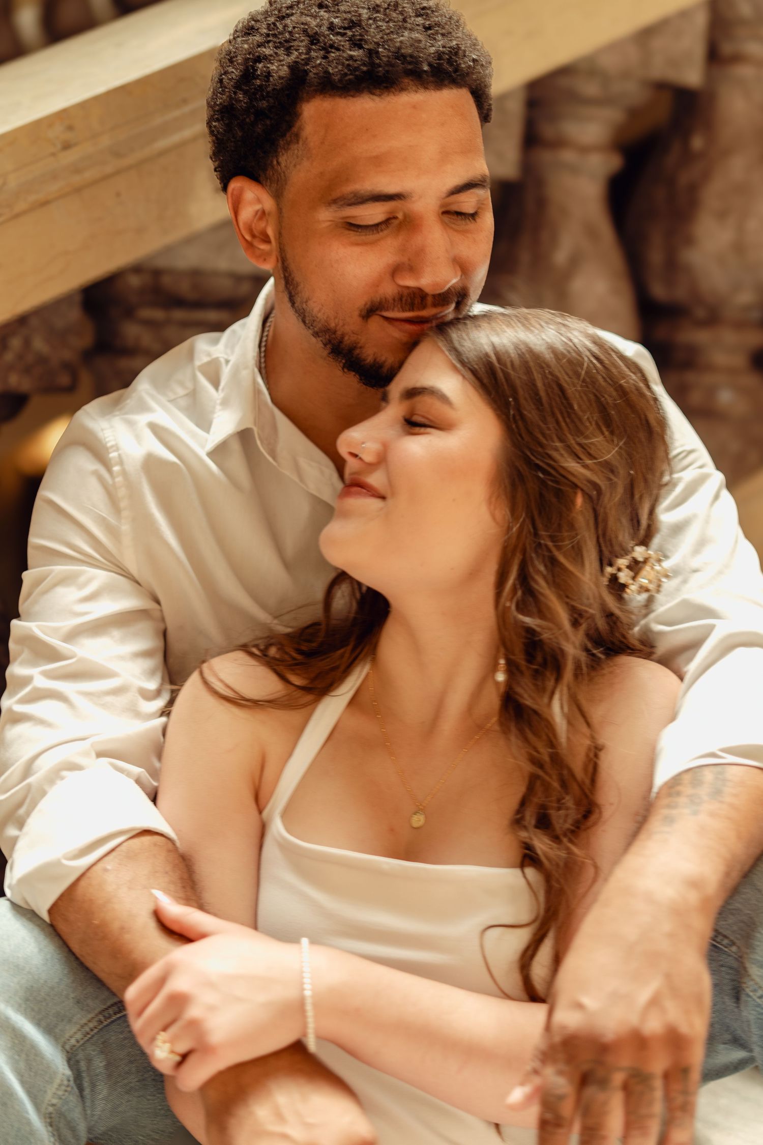 A romantic interracial couple sharing a tender moment, woman in white dress leaning back against man in white shirt.