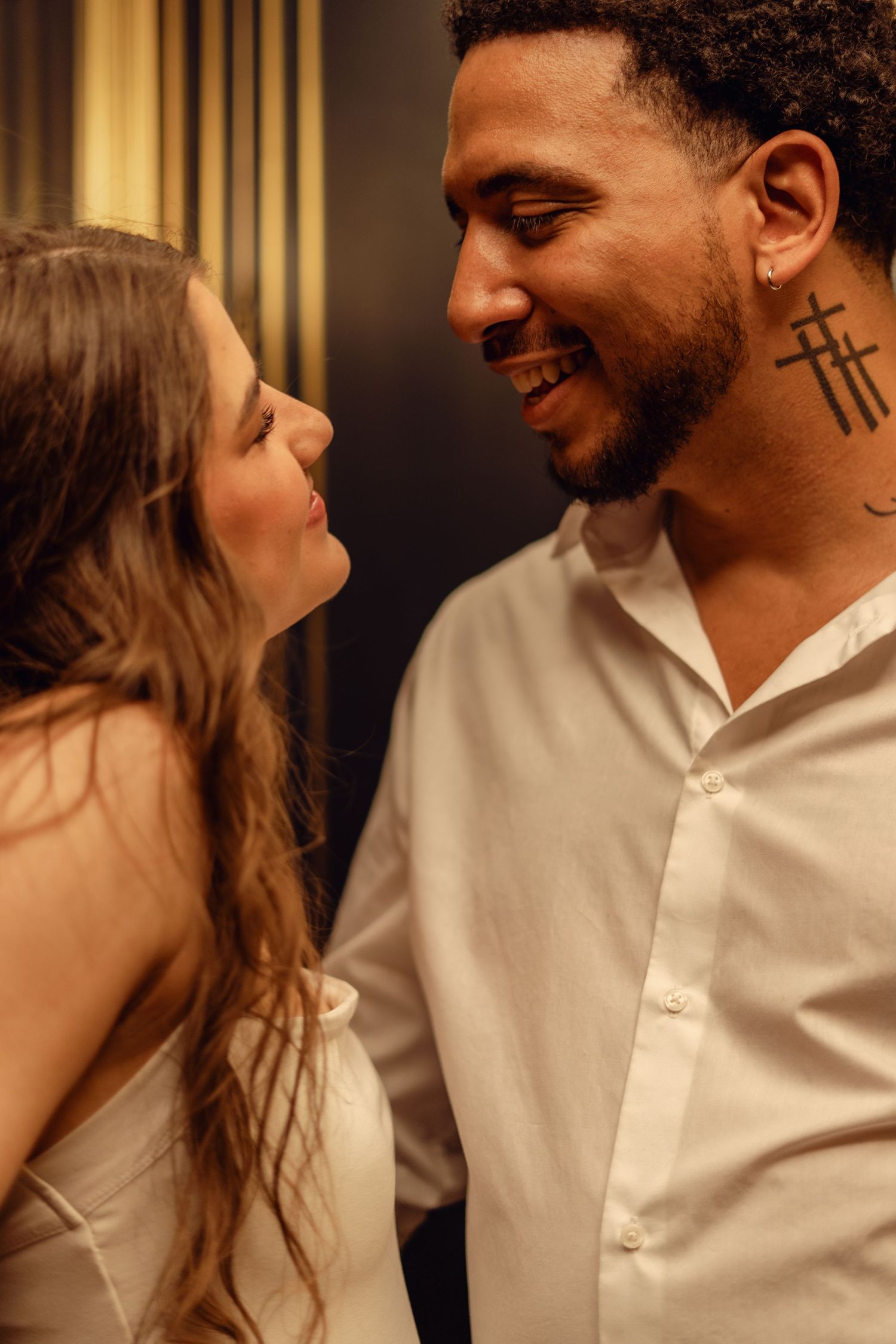 Romantic interracial couple gazing at each other, man with cross neck tattoo in white shirt, warm golden light.