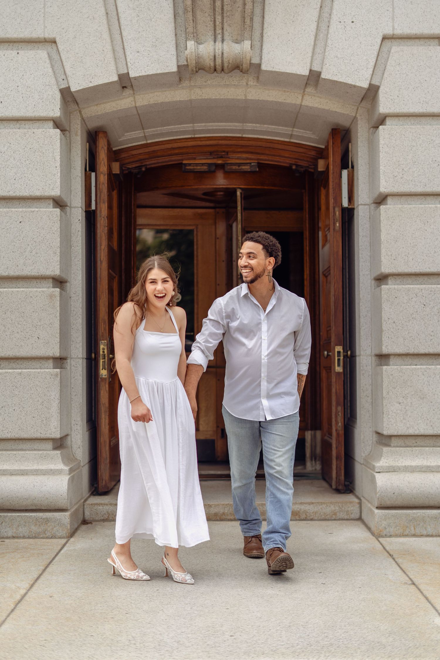 Happy couple holding hands walking out of elegant stone building, woman in white dress, man in casual shirt and jeans.