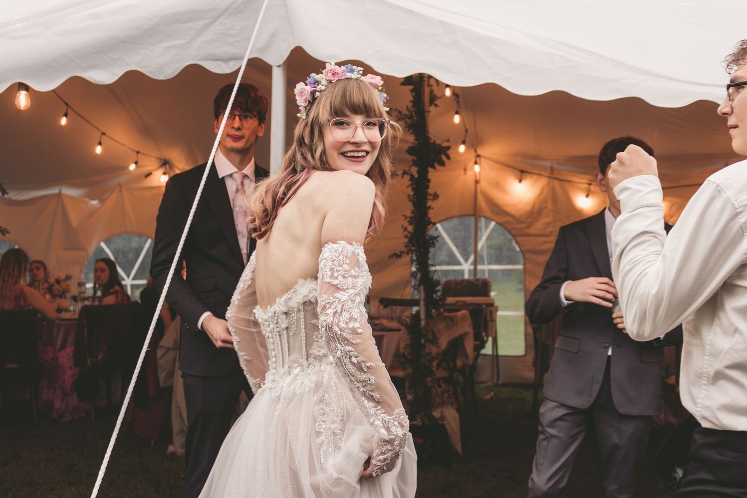 Smiling bride in lace wedding dress with floral crown dancing under string lights at elegant tent reception.