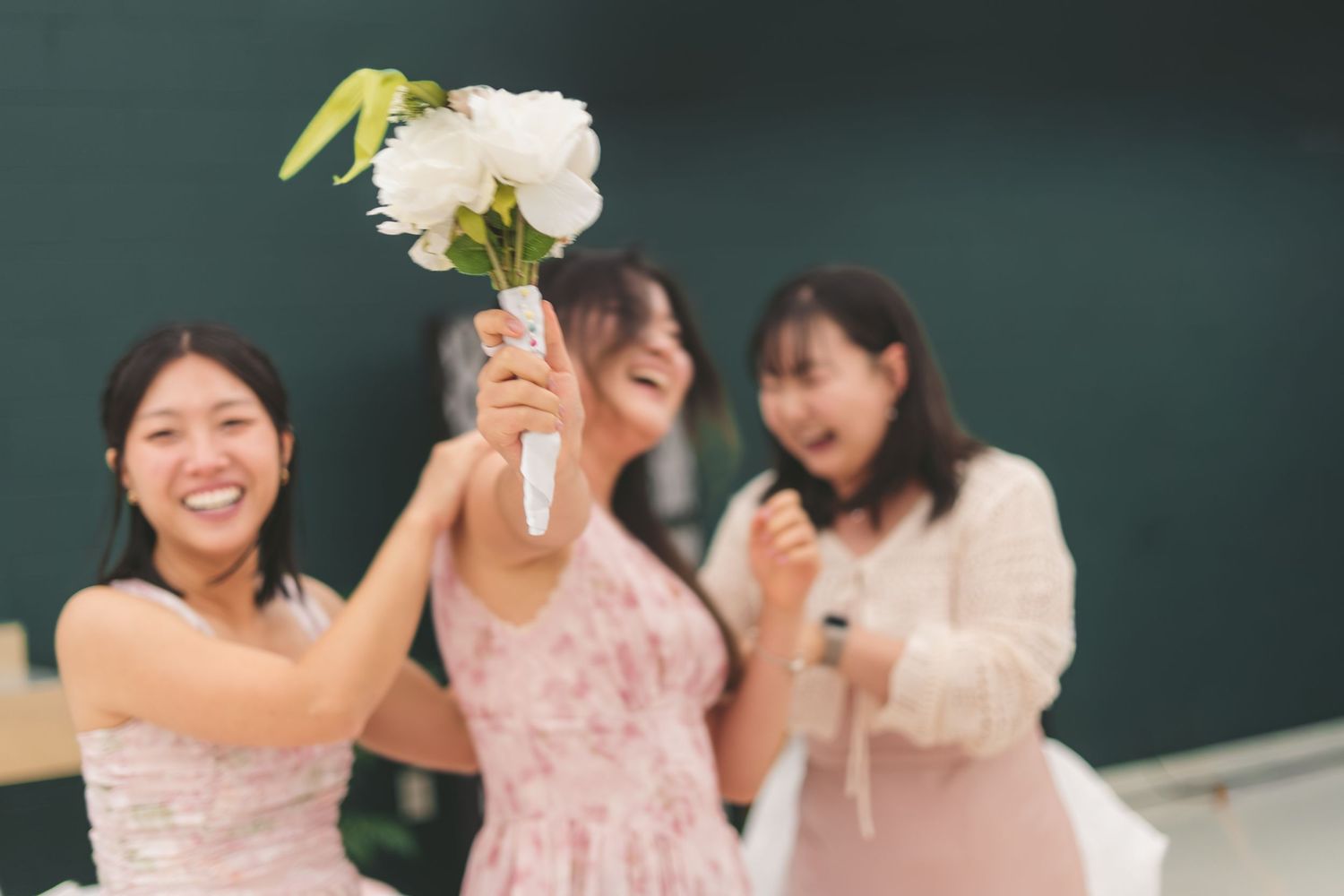Three young women celebrating with white peony bouquet, smiling and laughing in festive pink dresses.