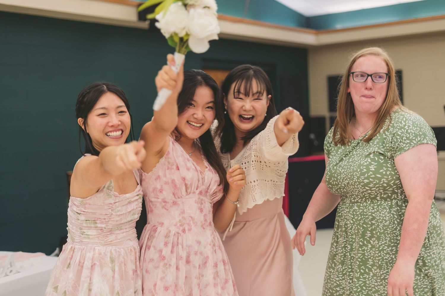 Four women smiling and celebrating at a wedding, one holding a bouquet, wearing floral and pastel dresses.