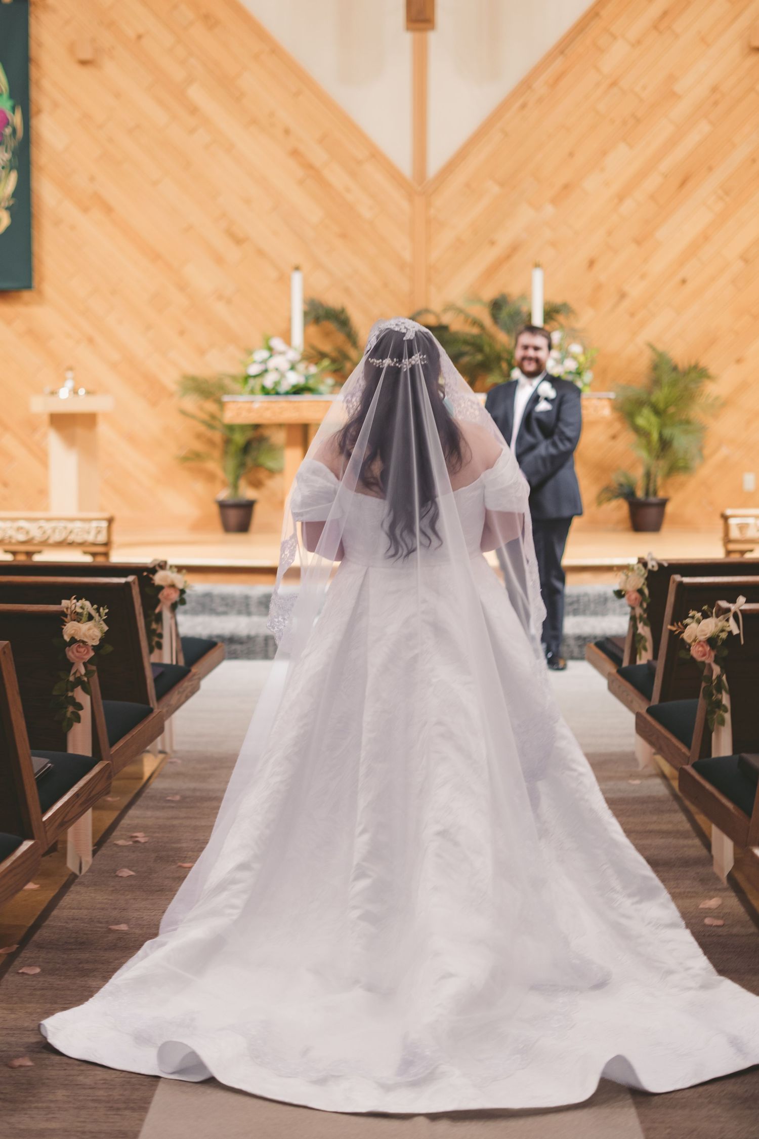 Bride walking down church aisle in white gown and veil toward groom at altar during wedding ceremony.