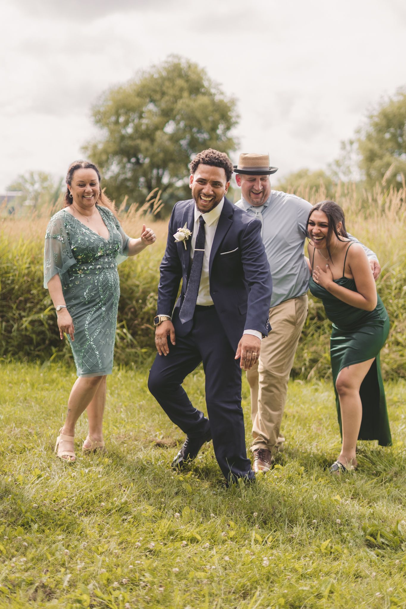 Groom in navy suit laughing with family on green grass at outdoor wedding celebration.