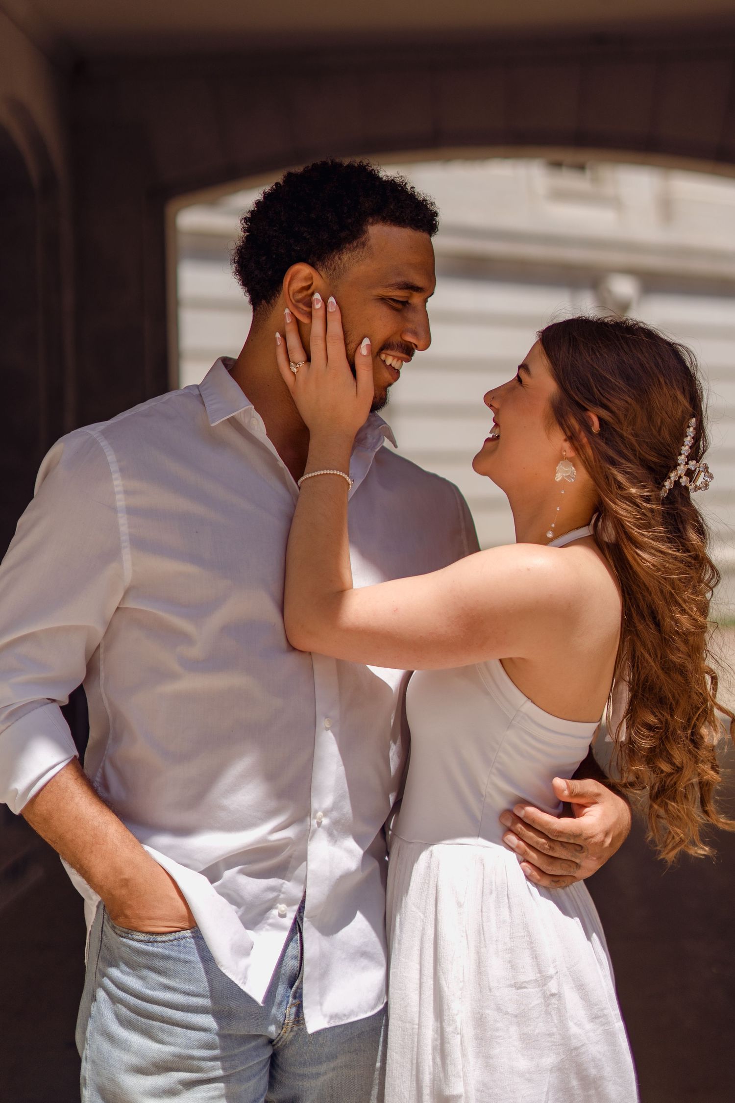 Interracial couple in white outfits sharing a joyful moment outdoors, bride touching groom's face tenderly.