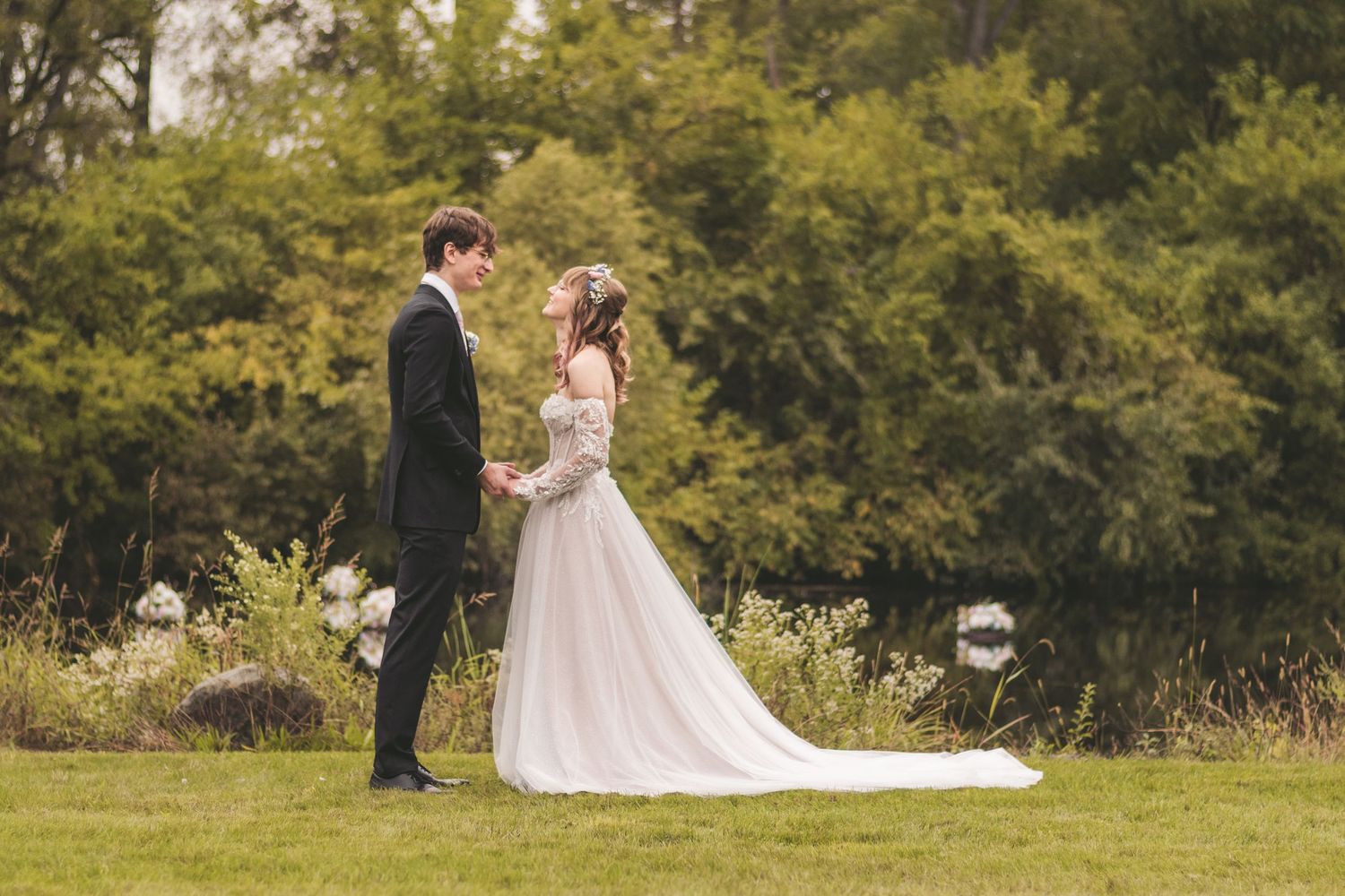 A bride and groom hold hands in a lush green garden, the bride wearing an elegant white gown with a flowing train.