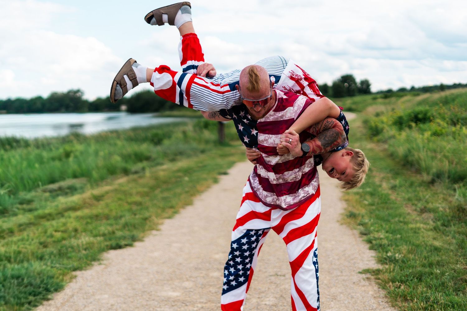 Two men in matching American flag outfits wrestling on a scenic path near a lake.