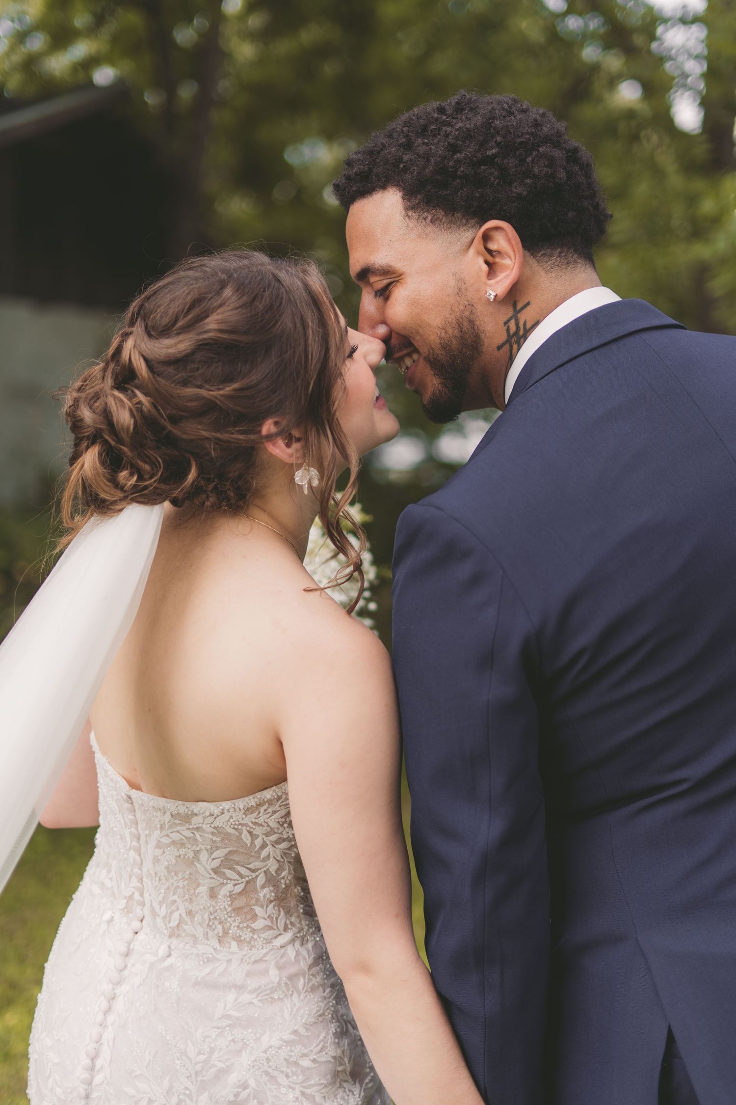 A bride and groom share an intimate moment, nearly kissing outdoors, she in a lace gown with veil, he in a navy suit.