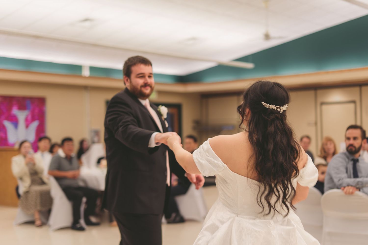 Bride and groom share their first dance at wedding reception, guests seated in background.