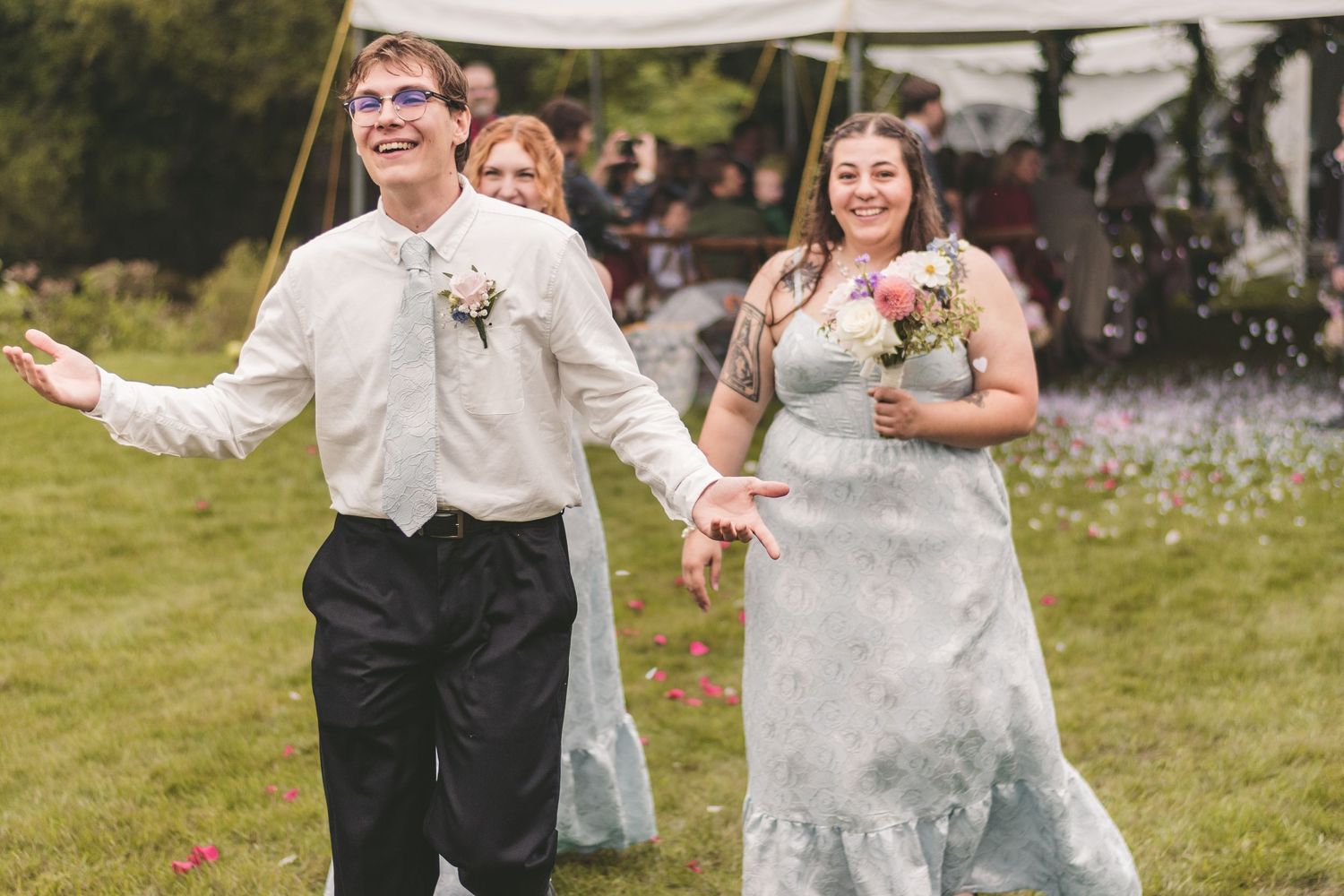 Joyful newlyweds running through confetti shower on green lawn outside white wedding tent.
