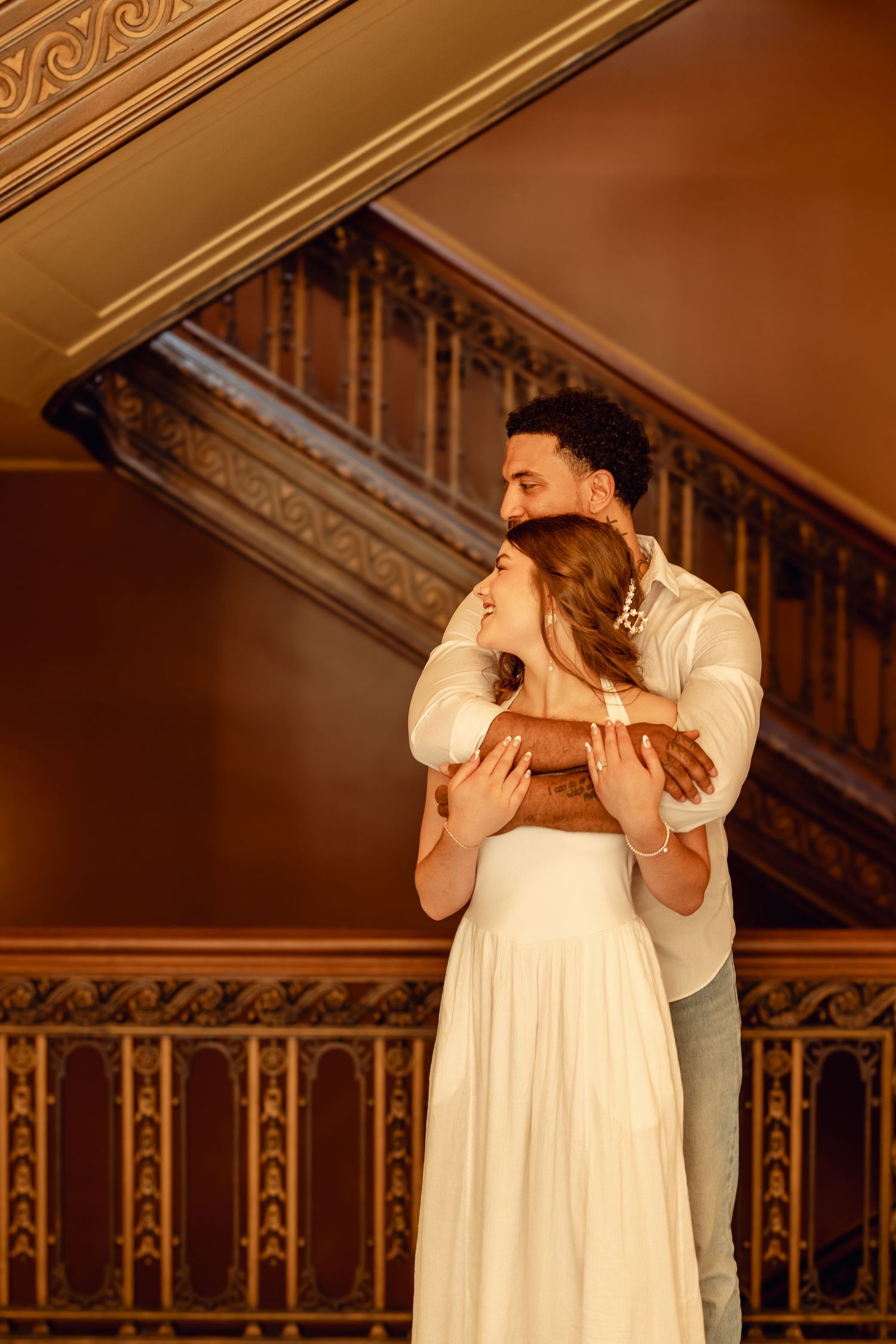 A couple embraces romantically on an ornate staircase, the woman in a white dress smiling warmly in a golden-lit venue.