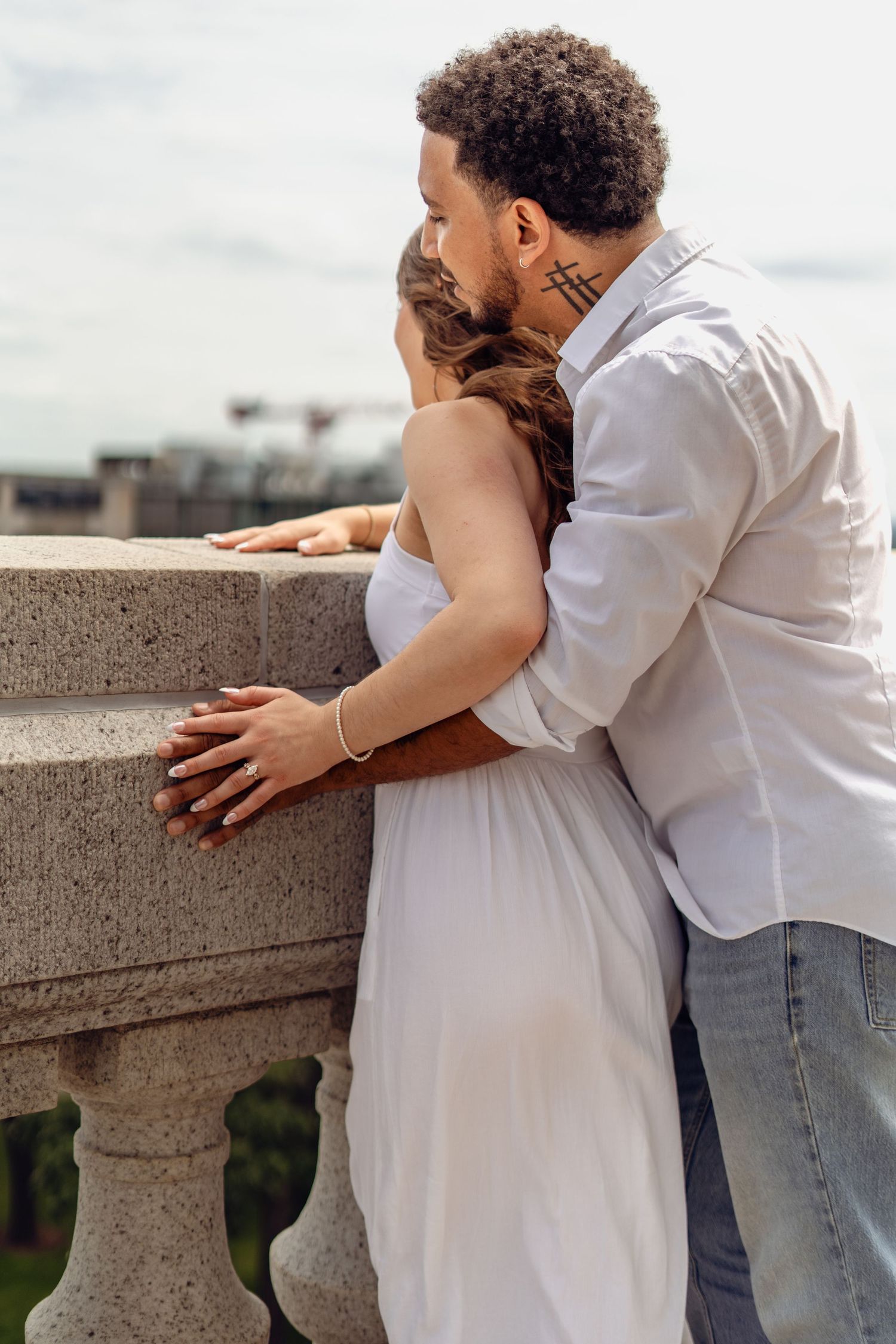 A loving couple embraces on a stone balcony, the man kissing the woman's head as they enjoy a scenic outdoor view.