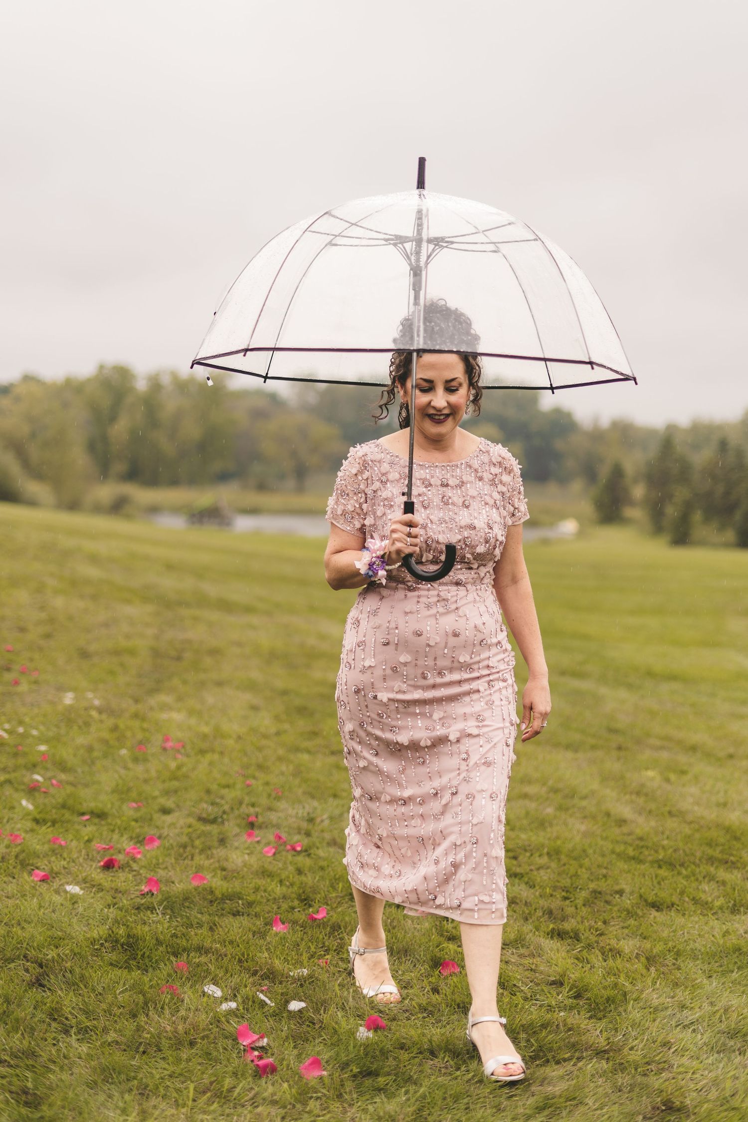 Woman in pink lace dress walking outdoors under a clear bubble umbrella on a cloudy day.