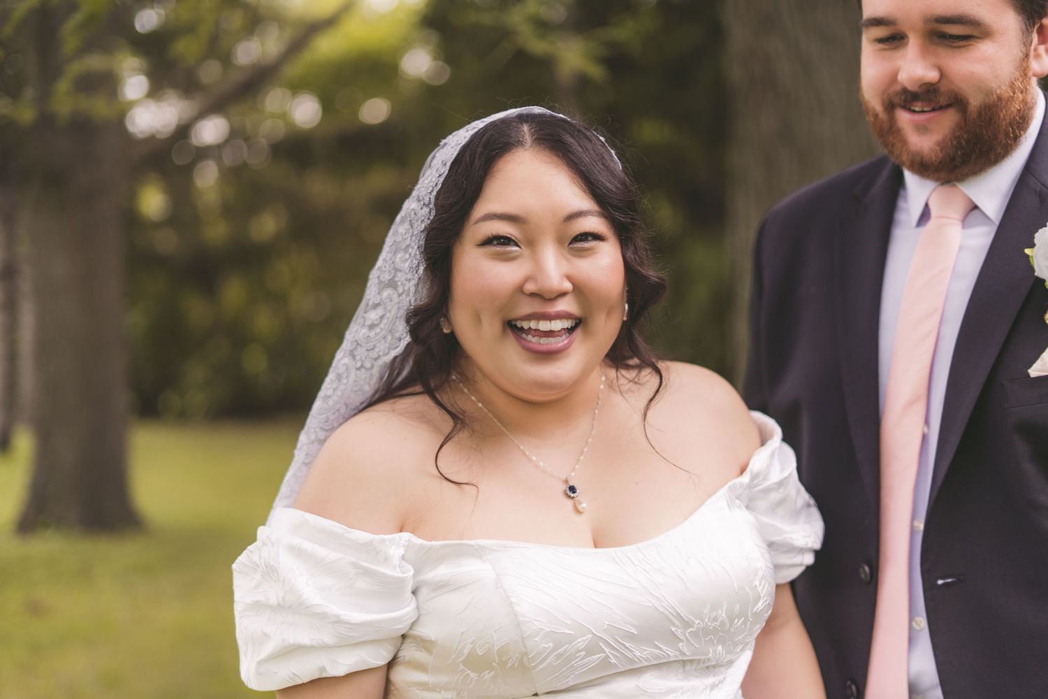 Smiling Asian bride in white off-shoulder wedding dress wearing a lace veil on her wedding day.