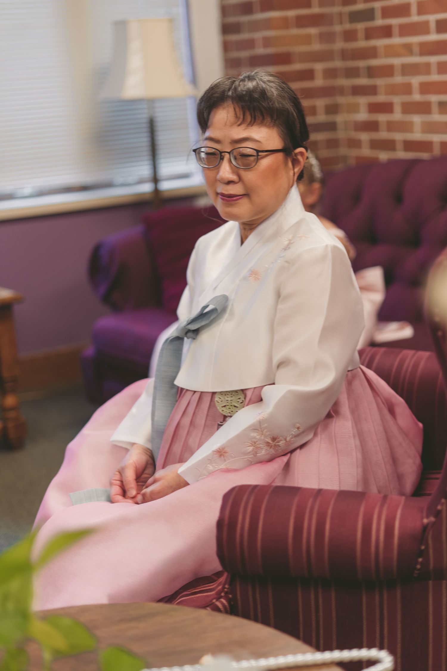 Elderly Korean woman in traditional white and pink hanbok sitting gracefully on a burgundy chair indoors.
