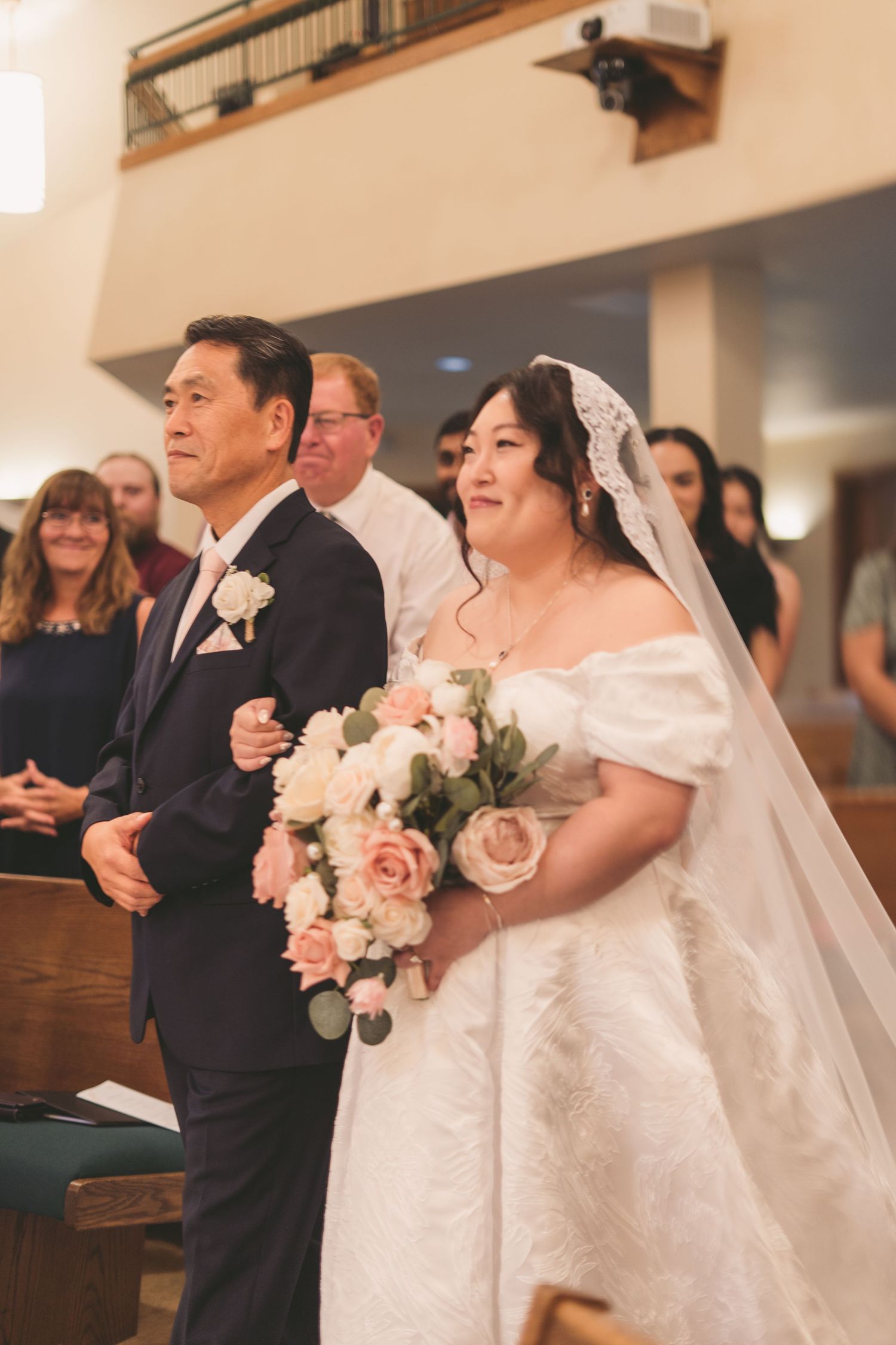 Bride in off-shoulder lace gown holding blush bouquet walks down church aisle with escort during wedding ceremony.