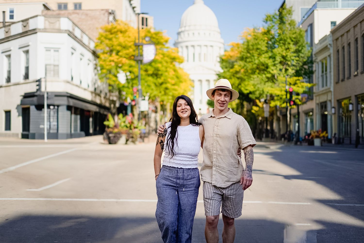 A couple walks down a city street with a white domed building visible in the background on a sunny autumn day.