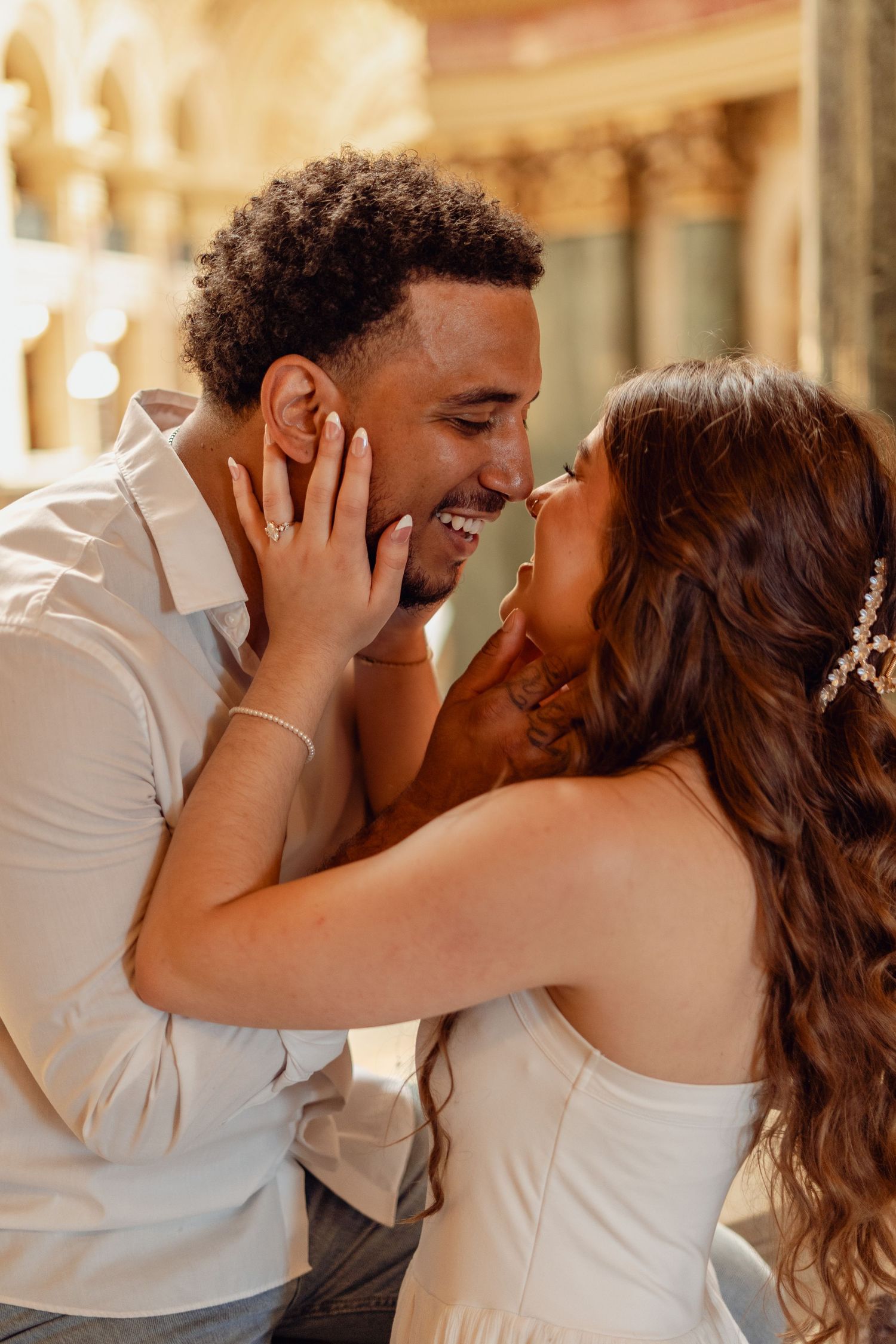 A joyful interracial couple shares a romantic moment, nearly kissing and laughing together in a sunlit architectural setting.