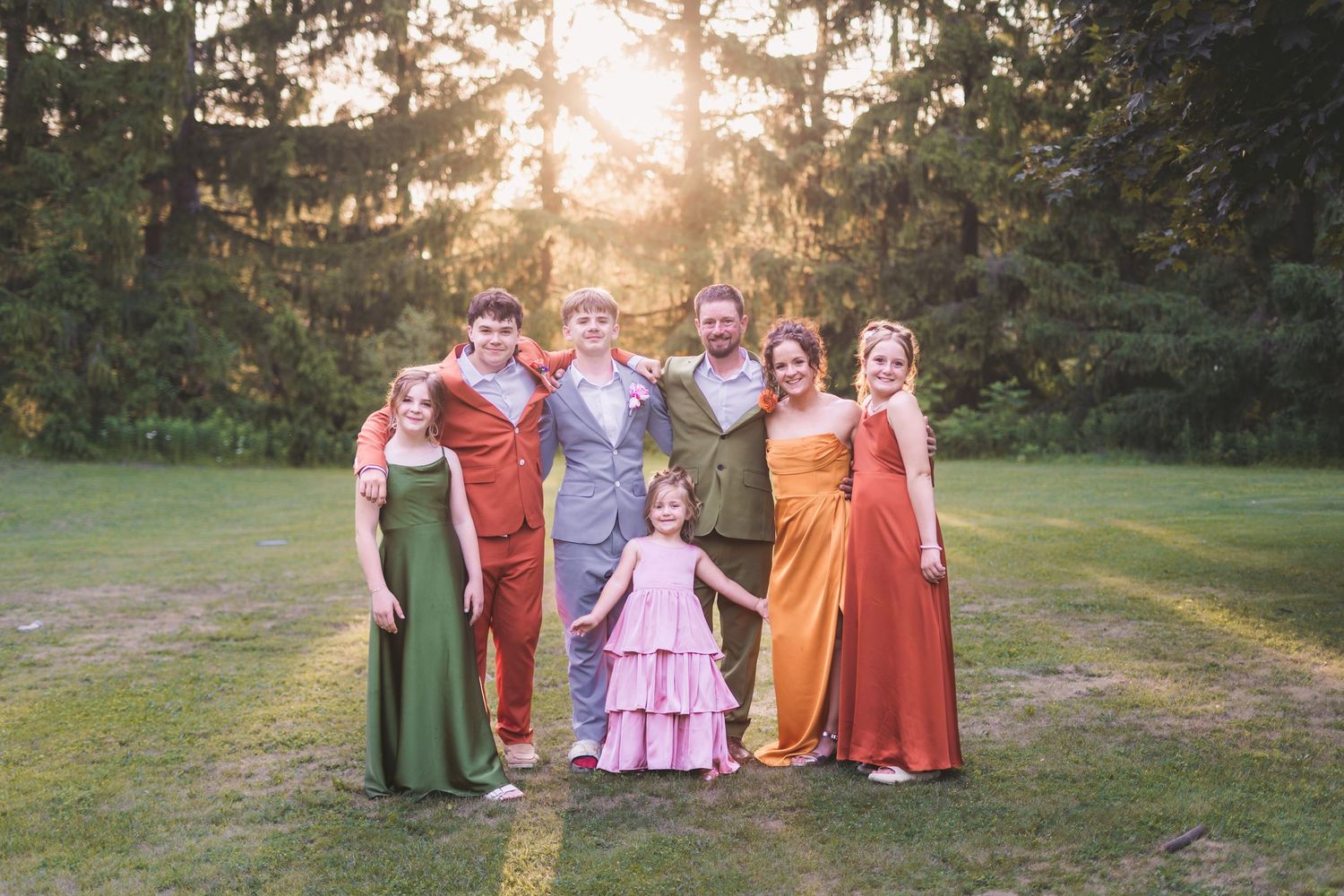 A joyful wedding party poses outdoors at sunset, wearing colorful gowns in green, red, orange, and pink amid lush trees.