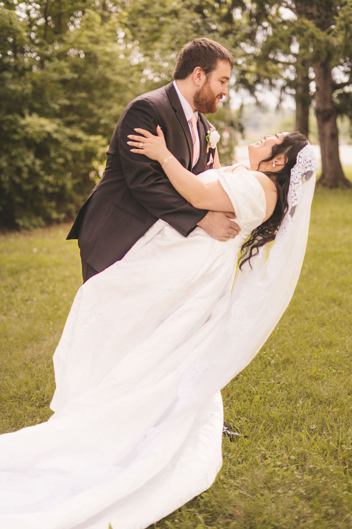 Groom dips bride in white wedding gown and veil outdoors on green grass, sharing a romantic moment.