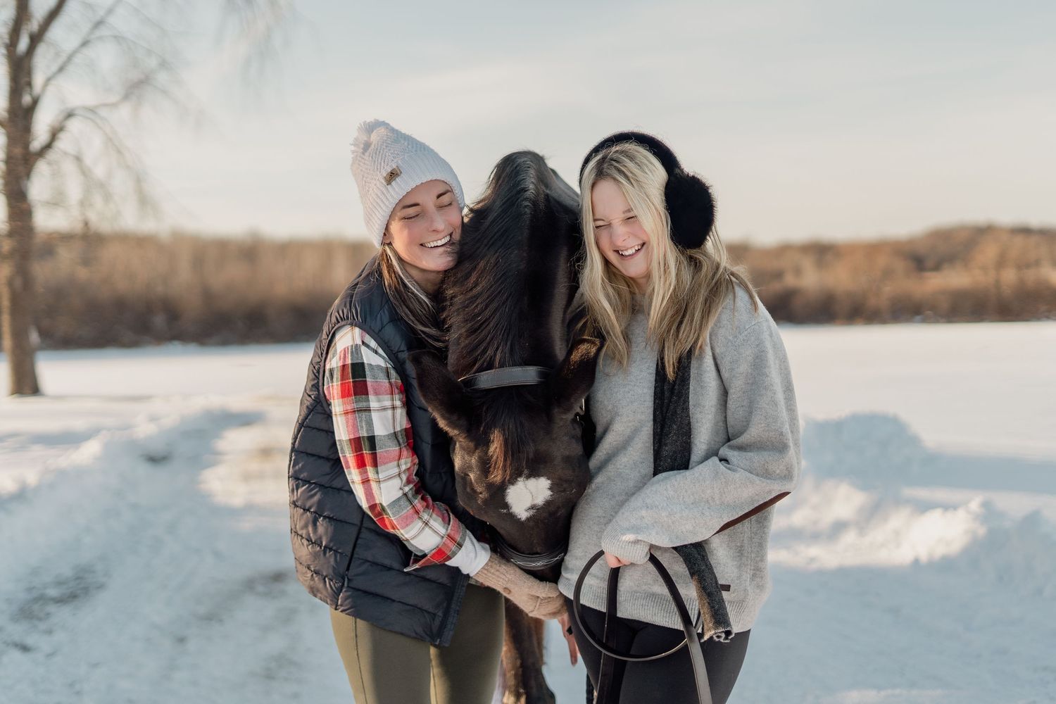 Two smiling women in winter clothing hug a dark horse outdoors in a snowy landscape on a sunny day.