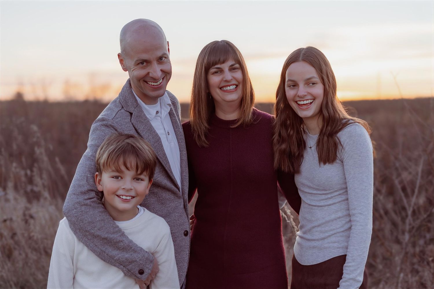 Happy family of four smiling together outdoors at sunset in a golden field.