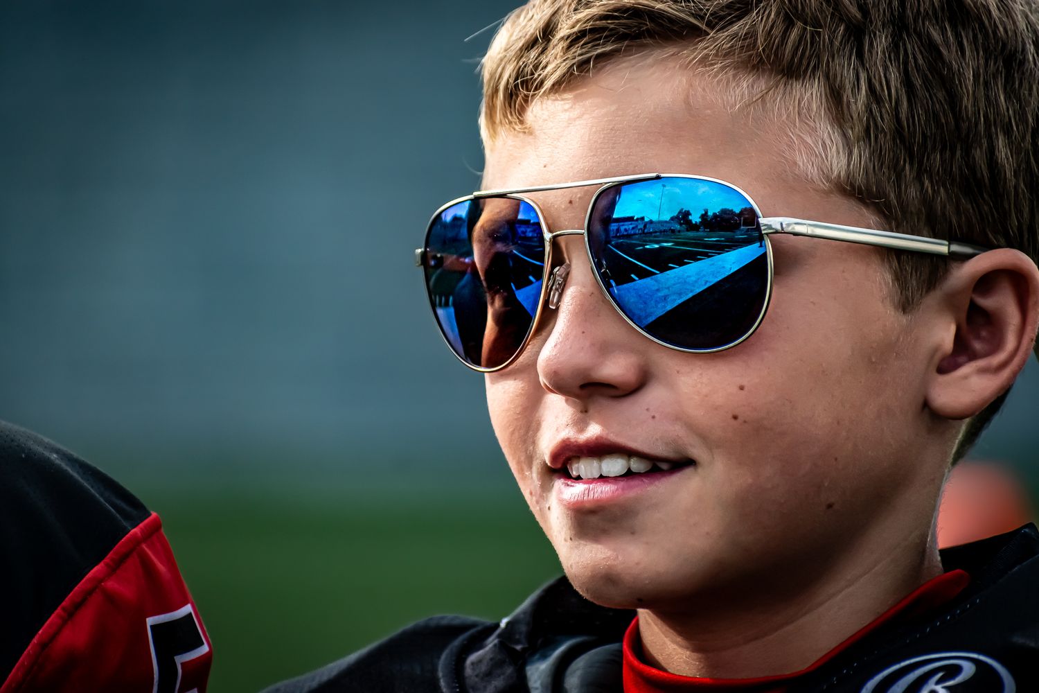 Smiling young boy wearing blue mirrored aviator sunglasses reflecting a racetrack, dressed in a racing jacket.