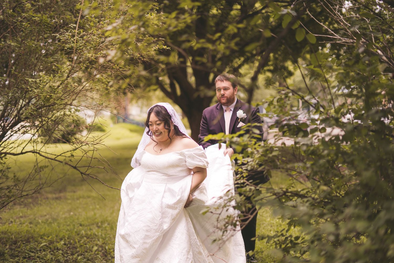 Bride in white gown and veil walks through lush green garden as groom follows behind on wedding day.